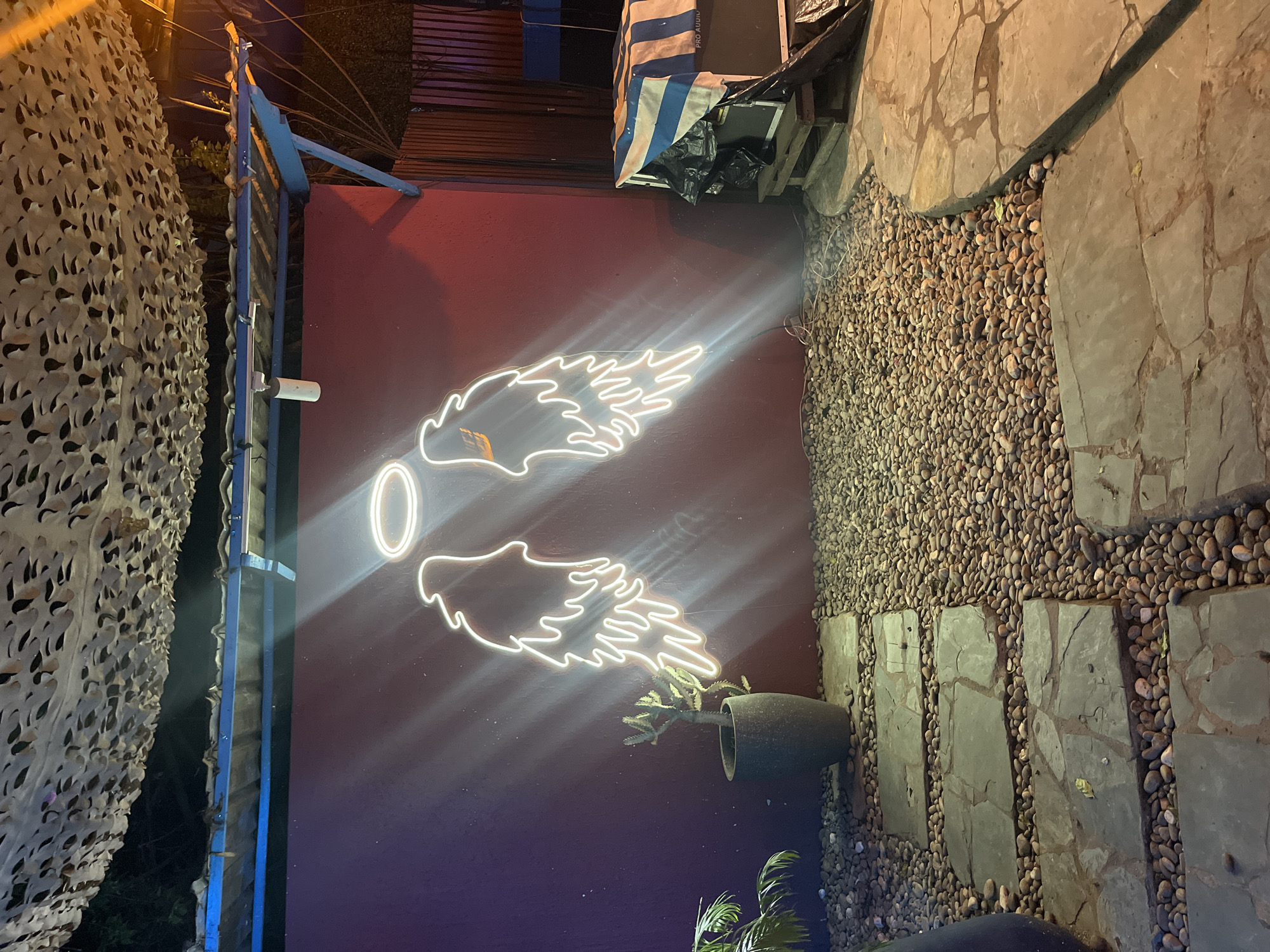 The image shows an outdoor scene at night in Accra, Ghana. The primary subjects are a neon wing art installation on a dark purple wall and a path made of stone slabs interspersed with small pebbles. The neon wings are white and emit a bright glow, with a distinct "O" shape positioned between them.
To the left of the wall, there is a blue metal structure and a camouflage-like netting. Further to the left and slightly behind, some foliage is visible, suggesting a natural element to the setting. To the right of the wall, the path leads towards a darker area, with more foliage and a potted plant visible near the wall. The overall atmosphere is illuminated by the neon sign and ambient lighting, creating a slightly mysterious and inviting feel.
The scene appears to be part of an outdoor space, possibly a lounge, bar, or a public area designed for visual appeal. The presence of stone and pebbles, along with the potted plant, indicates a landscaped environment. There is no clear activity or event taking place; it is a static scene. The time of day is definitively night due to the darkness and the reliance on artificial light sources, specifically the neon sign. The location cues (Accra, Ghana) combined with the warm night lighting suggest a tropical climate. There is no visible text in the image.