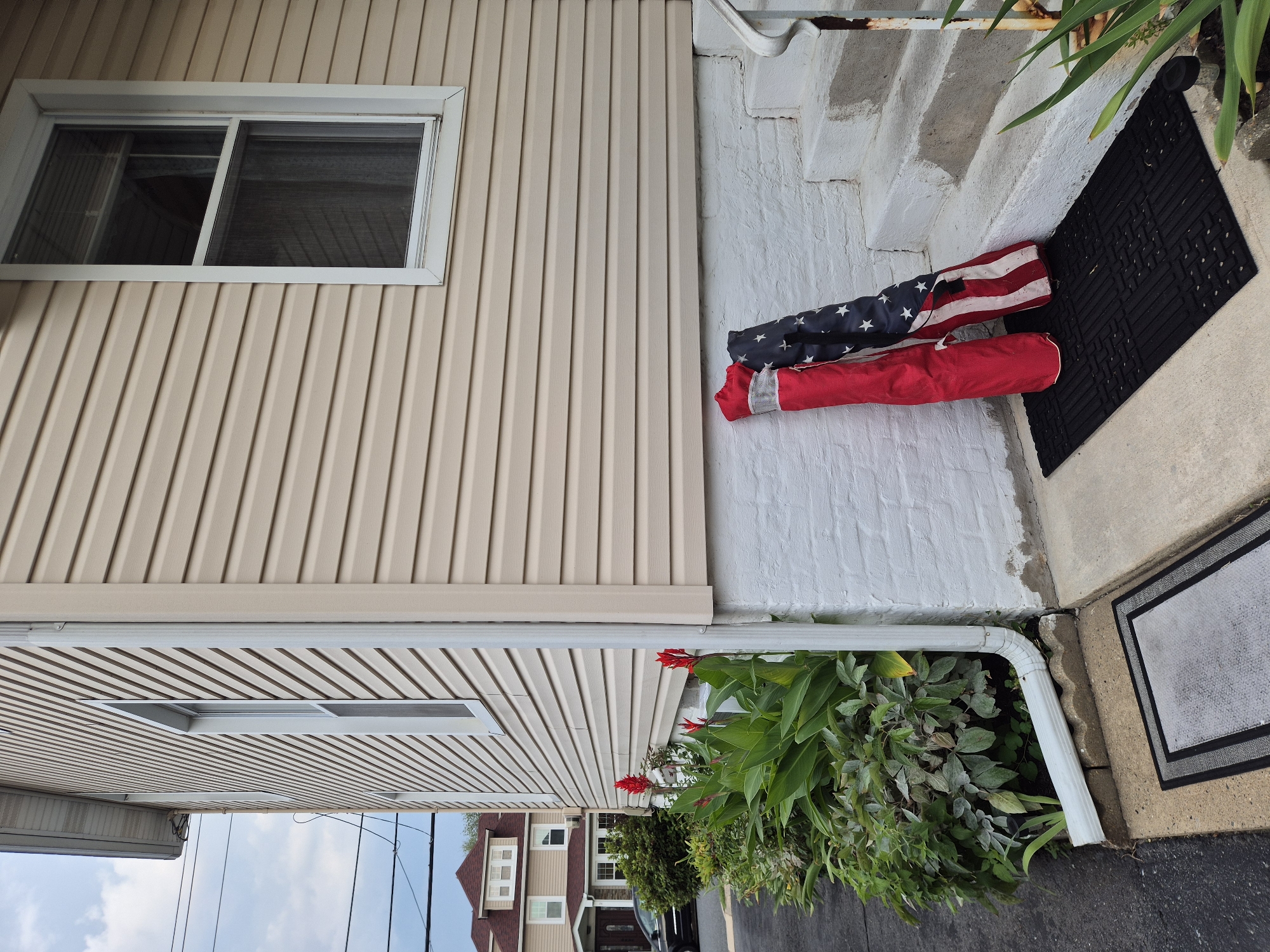 The image shows the exterior of a two-story house in South River, United States of America. The house has light beige vinyl siding and white trim around its windows. A white downspout runs vertically along the corner of the house, feeding into a horizontal drainpipe. In front of the house, a row of green plants with red flowers, possibly canna lilies, are visible. A black car is parked on the asphalt driveway to the left of the plants.
In the foreground, two rolled-up items lean against the white-painted brick foundation of the house. One is predominantly red with a silver reflective stripe, and the other is an American flag with red and white stripes and a field of white stars on a blue background. Both are standing upright on a black rubber doormat. Concrete steps lead up to a doorway, with a rusty metal handrail on the right side of the steps. A gray doormat is situated on the concrete sidewalk to the left of the steps. The sky above is partly cloudy.