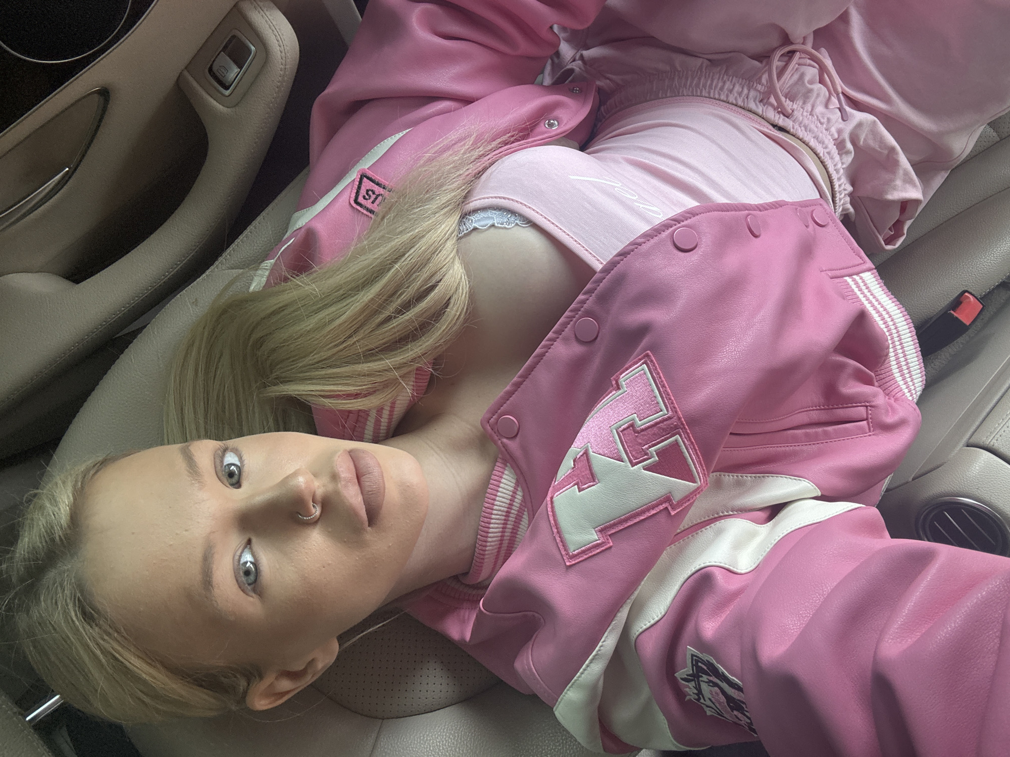 A young woman with blonde hair, wearing a pink jacket, a pink tank top, and pink pants, is sitting in the passenger seat of a car. She is looking directly at the camera with a neutral expression. The interior of the car is visible, with leather seats and a car door. The scene is indoors, likely in Saint Petersburg, Russia, given the details of the car's interior which are common in that region.