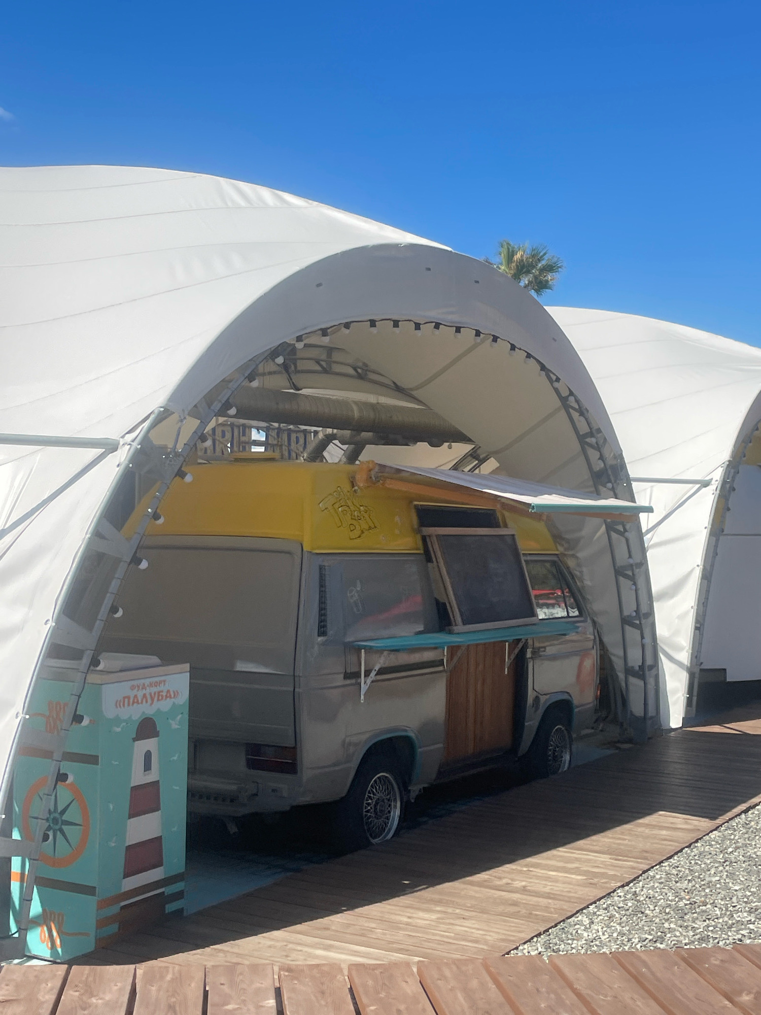 A vintage Volkswagen van, painted yellow on top and silver below, is parked under a white arched tent structure in Sochi, Russia. The van appears to be a food truck, with a serving window open and a blackboard displayed. Adjacent to the van is a teal-colored stand decorated with a lighthouse and a compass rose, with Russian text that translates to "Food Court 'Paluba'". The ground is covered with wooden planks and gravel. A palm tree is visible in the distance against a clear blue sky. There are no people visible in the image.