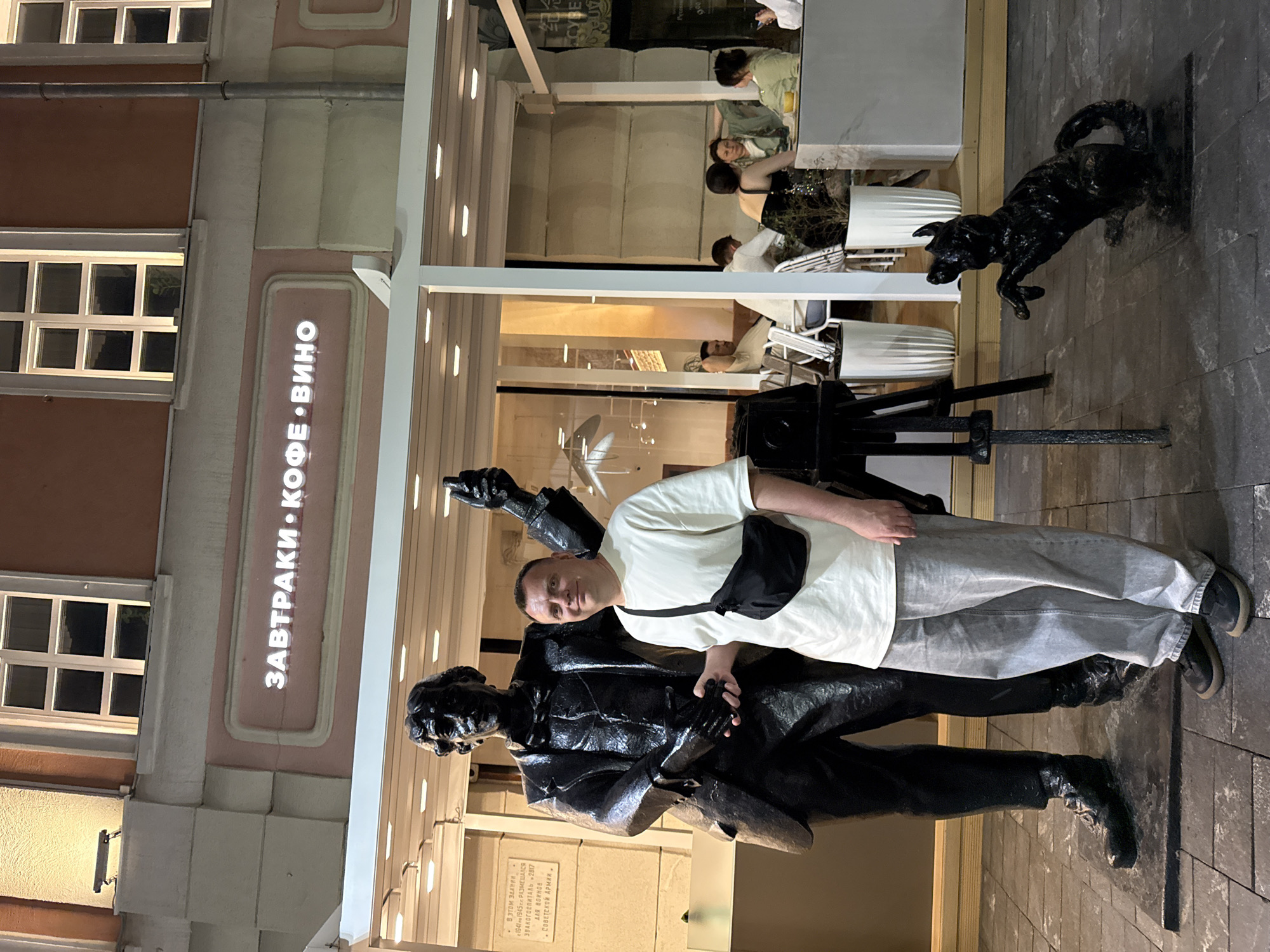 The image captures a man posing next to a statue of a scientist and his dog in Nizhny Novgorod, Russia. The man is wearing a white t-shirt, grey pants, a black shoulder bag, and black shoes. He is standing on a tiled sidewalk, leaning slightly against the statue. The statue is bronze or a similar dark material, depicting a man holding an object and a dog standing alert beside him. Behind the statue is a building with a sign in Russian that reads "ZAVTRAKI * KOFE * VINO", translated as "Breakfast * Coffee * Wine". In the background, there is a seating area with several people at tables, suggesting an outdoor café or restaurant setting. The building's exterior has a combination of pink and white colors, with windows on the upper floors. The overall lighting is soft and suggests an evening setting.