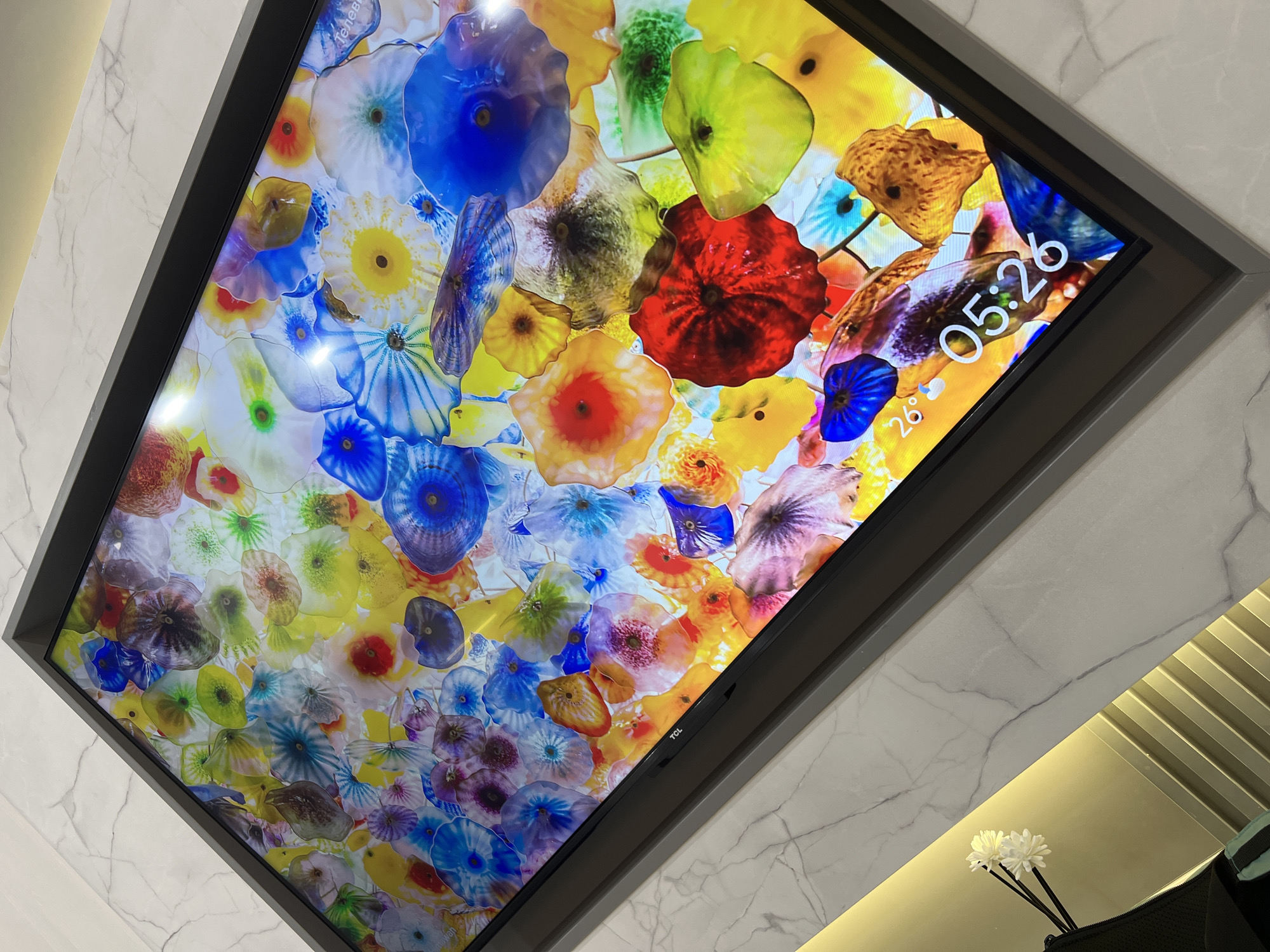 The image shows a close-up view of a modern television screen displaying a vibrant, abstract pattern of colorful glass or floral shapes. The screen is framed by a dark bezel and sits on a light-colored, marble-patterned surface. In the lower left corner of the screen, a digital clock displays "05:26" and a temperature of "26°". The surrounding environment includes a wall with a marble-like finish and subtle lighting, with a glimpse of a decorative object or flower arrangement in the lower left of the frame. The scene appears to be indoors, in a contemporary setting. No specific location in Bac Nha Trang Ward, Vietnam can be determined from the provided image.