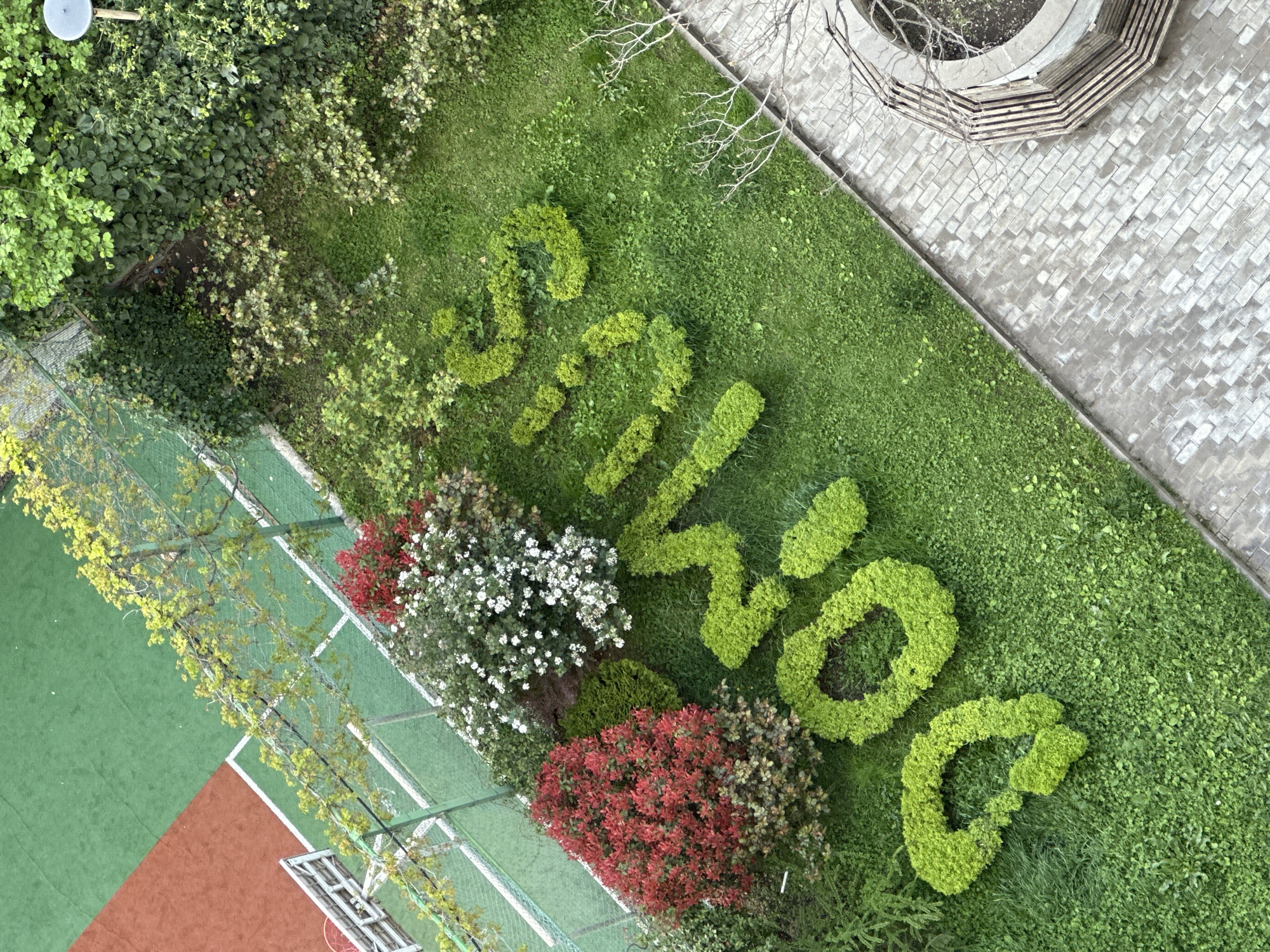 An aerial view of a green lawn in Tbilisi, Georgia, shows "Sakartvelo" spelled out in shrubbery. Red, white, and green shrubbery lie adjacent to the name. To the bottom left is a tennis court; to the top right, a paved area with a circular bench around a tree. Other green foliage surrounds the manicured lawn.