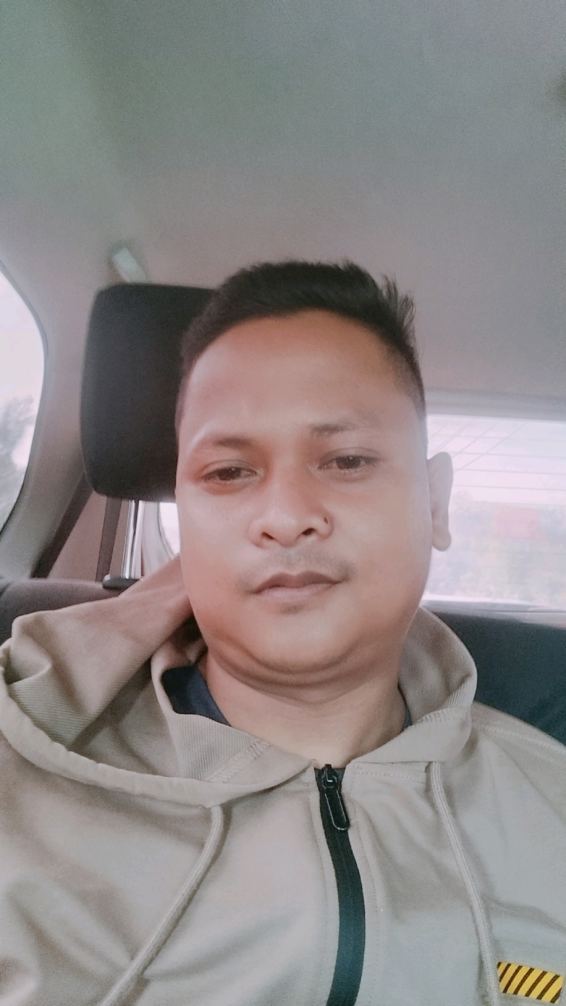 The image is a selfie of a man in a car, likely taken in Bintangpermata, Indonesia. The man is wearing a tan hooded jacket with a black zipper. He has short, dark hair and is looking directly at the camera. The background shows the interior of a car, with a headrest visible behind him and a window with a blurred outdoor scene beyond. The lighting appears to be natural, coming from the front and possibly the side. The overall impression is a casual, candid shot.