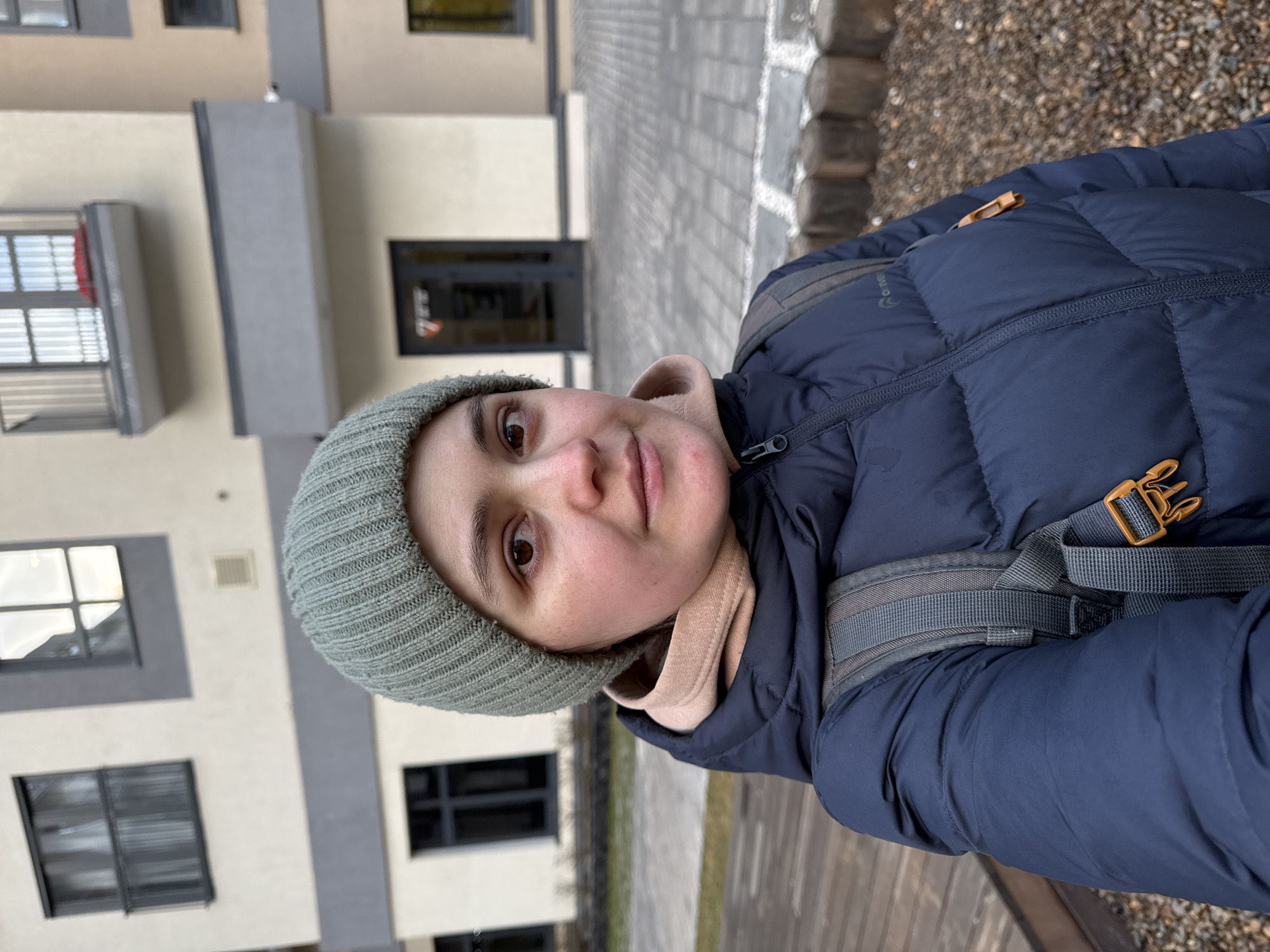 A young adult woman is captured in a close-up selfie outdoors, looking directly into the camera with a neutral expression. She has light brown eyes and is wearing a warm ensemble suitable for cool weather. Her attire consists of a dark blue puffer jacket with horizontal quilting, a light beige hooded sweatshirt underneath, and a sage green ribbed knitted beanie. Grey backpack straps with an orange/brown buckle are visible over her right shoulder. The word "ENVY" along with a circular logo is discernible on the left side of her jacket.
The setting appears to be an urban or suburban environment in Izhevsk, Russia. Behind her, a modern, light-colored building with grey architectural accents and dark-framed windows is visible. The ground is a mix of light-colored paving stones and a textured area, possibly gravel or wood chips. The lighting suggests an overcast daytime.