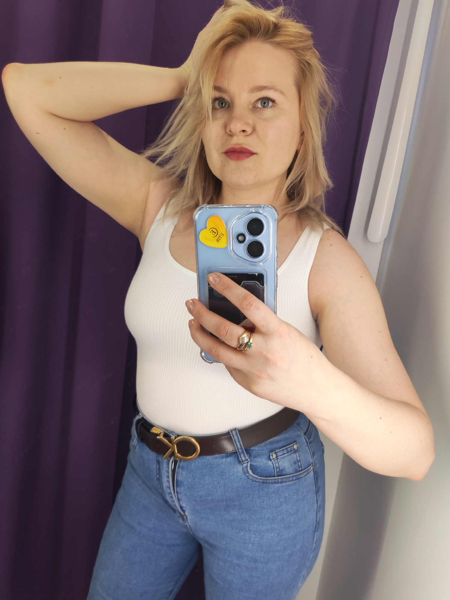 A woman is taking a selfie in what appears to be a fitting room. She has blond hair, blue eyes, and is wearing a white tank top, blue jeans with a brown belt, and a gold ring with a green stone. Her left arm is raised, and her right hand holds a blue phone with a yellow heart sticker. The background consists of a purple curtain and a white wall. The location is not identifiable as Izhevsk, Russia, from the image.