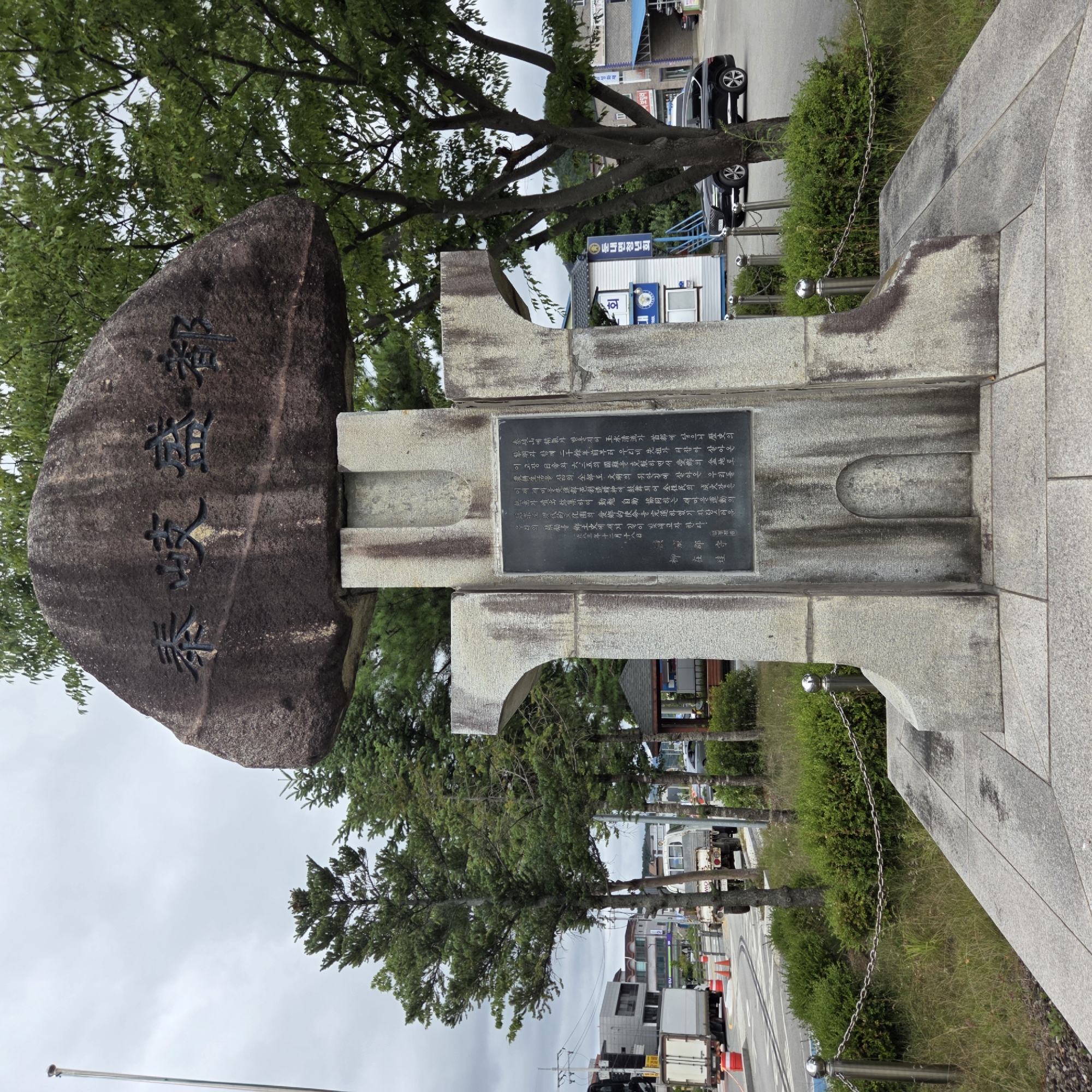 This is a medium-shot, eye-level photograph of a monument with Korean inscriptions, located in Dunnae-myeon, South Korea. The monument is made of stone, with a large, dark, irregular rock at the top inscribed with Korean characters. Below this rock is a rectangular stone base with a black plaque also inscribed with Korean text. The monument is situated on a paved area, surrounded by green grass and manicured bushes.
In the background, a street can be seen with traffic, including a white truck and a black SUV. There are also several trees, power lines, and buildings visible. The sky is overcast. The overall impression is that of a civic monument in an outdoor urban or semi-urban setting.
