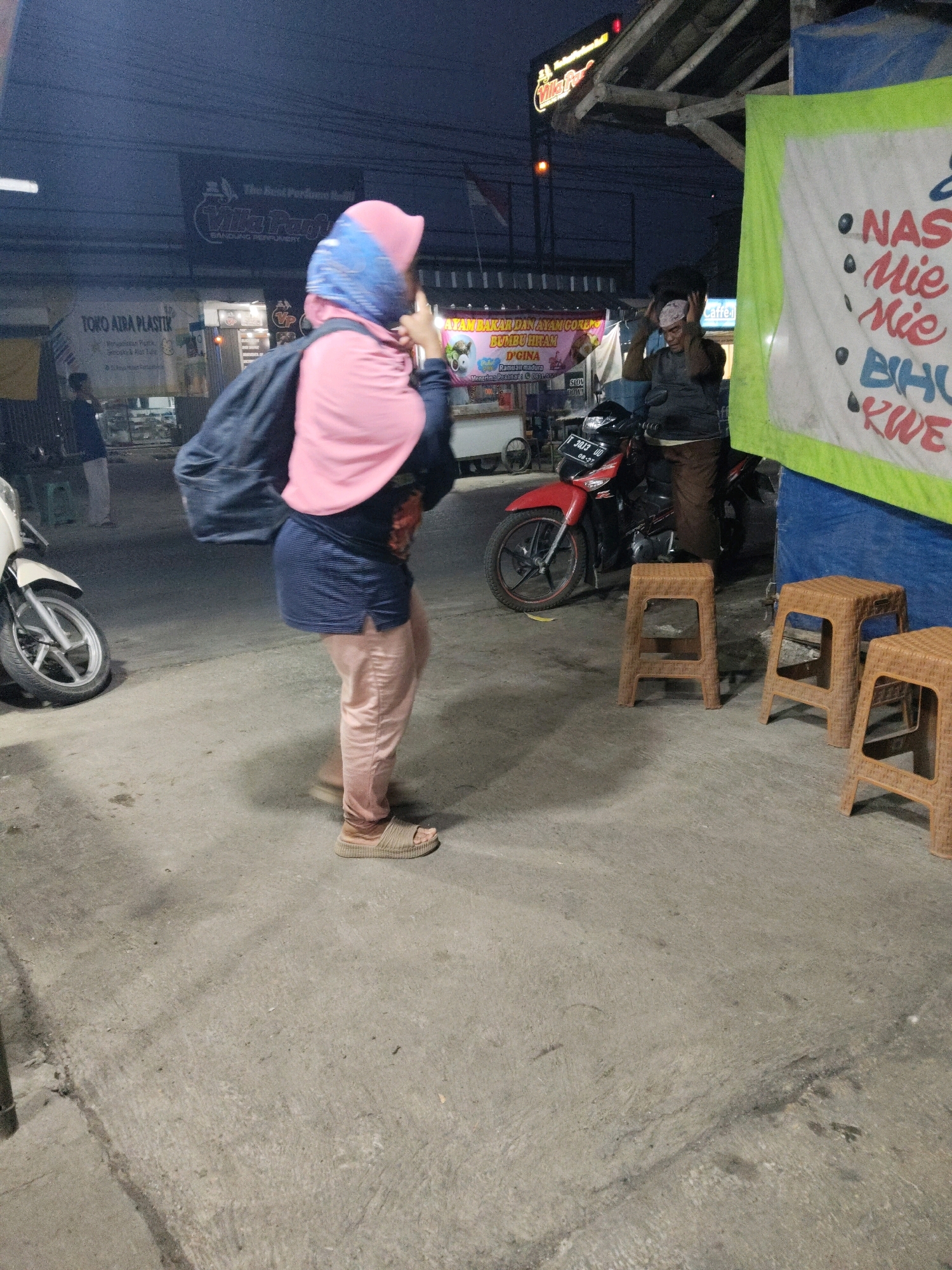 A woman wearing a pink hijab, dark blue top, and light pink pants walks on a concrete sidewalk at night. She carries a dark backpack. In the background, a red motorcycle is parked, and a man in a brown garment and white cap stands near it. A banner with green trim hangs to the right, displaying text in red and blue that appears to list food items. Another banner, partially visible on the left, has text in black and white. The setting seems to be an outdoor market or street food stall area. The lighting suggests it is evening or nighttime.