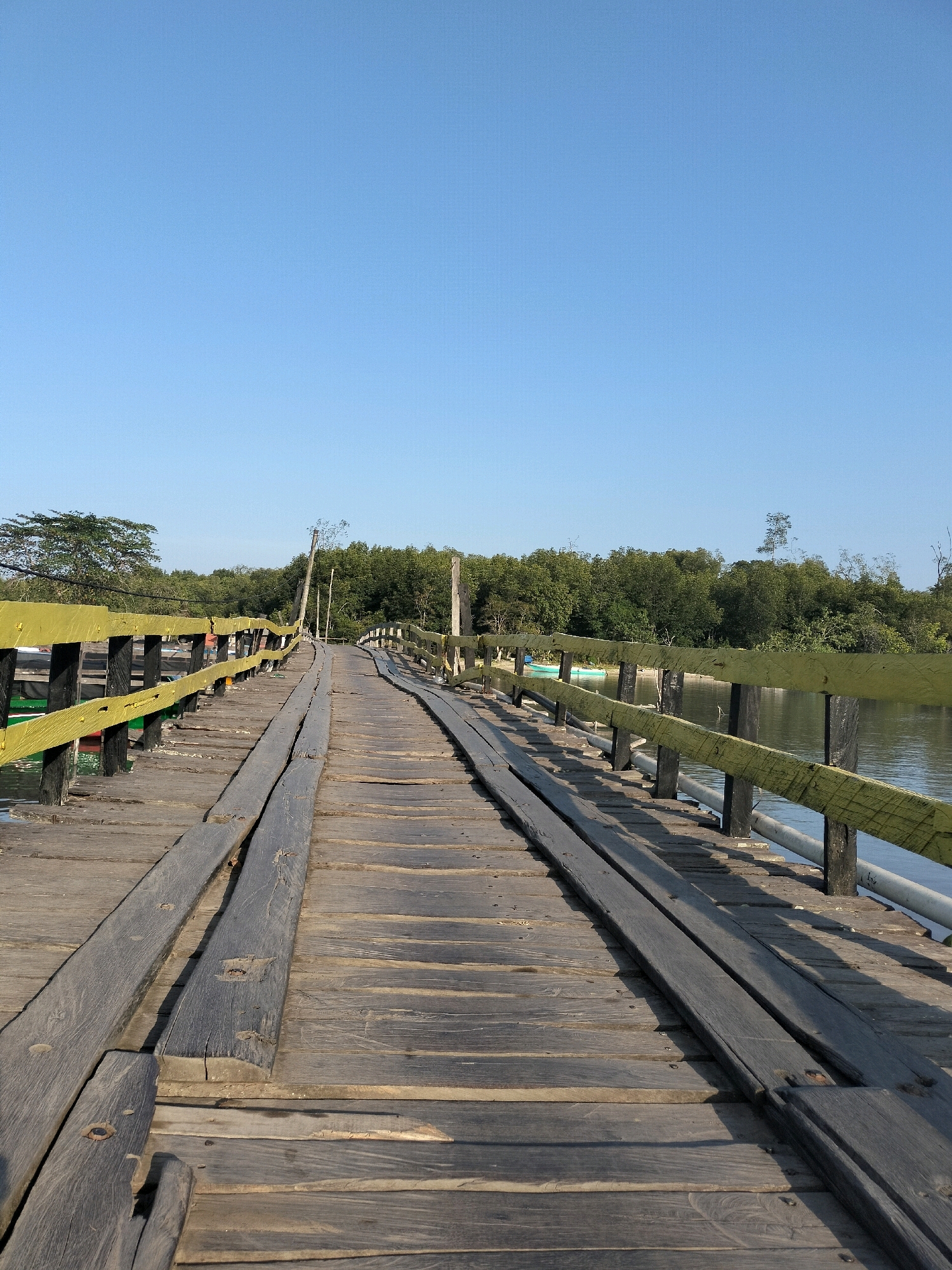 This image depicts a weathered wooden bridge stretching over a calm body of water. The bridge is constructed from aged, grey planks, some of which appear worn and slightly raised, creating a textured surface. A bright yellow railing with black posts lines the sides of the bridge, contrasting with the darker wood. Beyond the railing, a tranquil river or canal is visible, its surface reflecting the clear blue sky. On the far bank of the water, a dense line of lush green trees forms a natural backdrop. The sky above is a vast expanse of uninterrupted, pale blue. The scene evokes a sense of peaceful isolation and natural beauty. The image is a vertical, full shot of the bridge. The location is identified as Bunati, Indonesia.