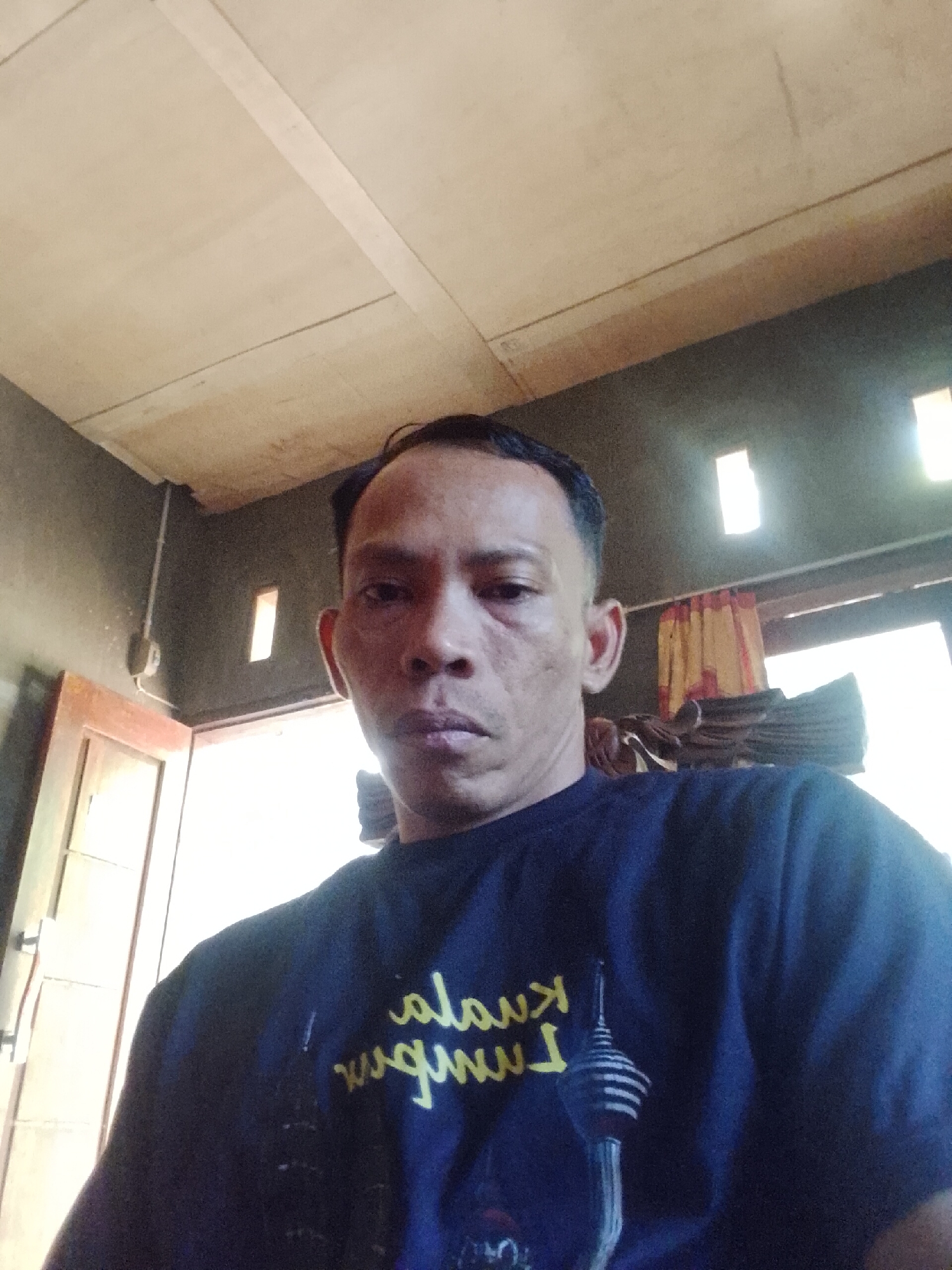 A man is pictured from the chest up, looking directly at the camera. He has short, dark hair and is wearing a dark blue t-shirt with yellow lettering and a graphic of a tower. The background appears to be an indoor setting with a wooden ceiling, a doorway on the left, and windows with curtains in the distance. The lighting suggests it is daytime. The location is Madukoro, Indonesia.