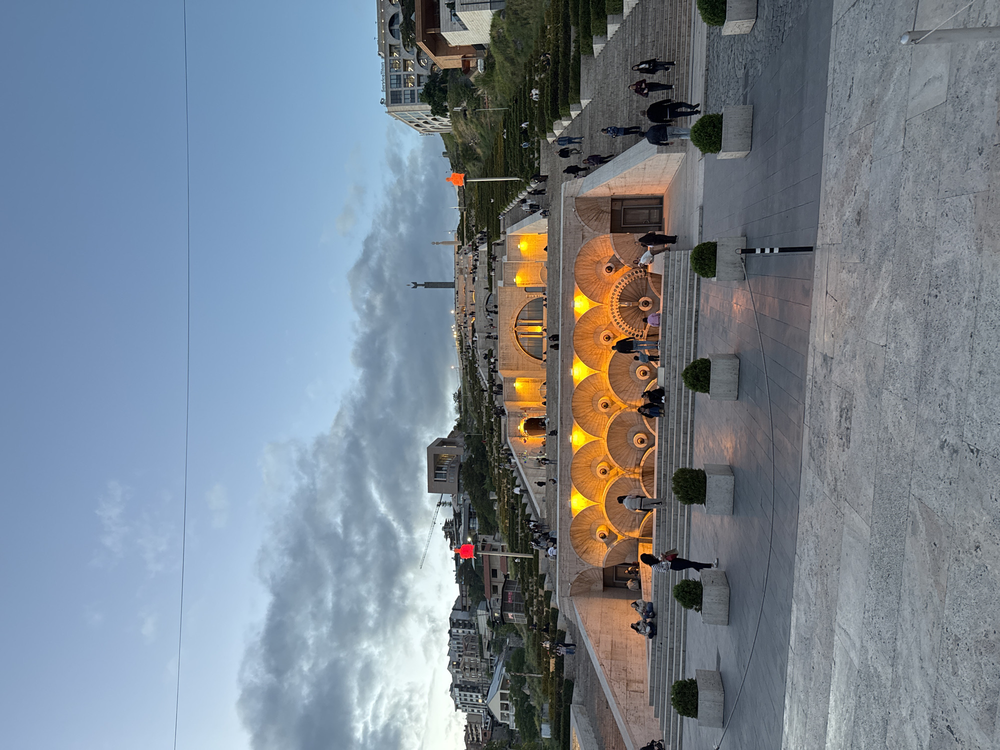 The image depicts the Cascade, a monumental stairway in Yerevan, Armenia. The structure features a series of illuminated fountains and levels. People are walking and sitting on the stairs. A red sculpture stands on one of the upper terraces. The sky transitions from blue to cloudy at dusk. Buildings are visible in the background. The foreground is a stone walkway.