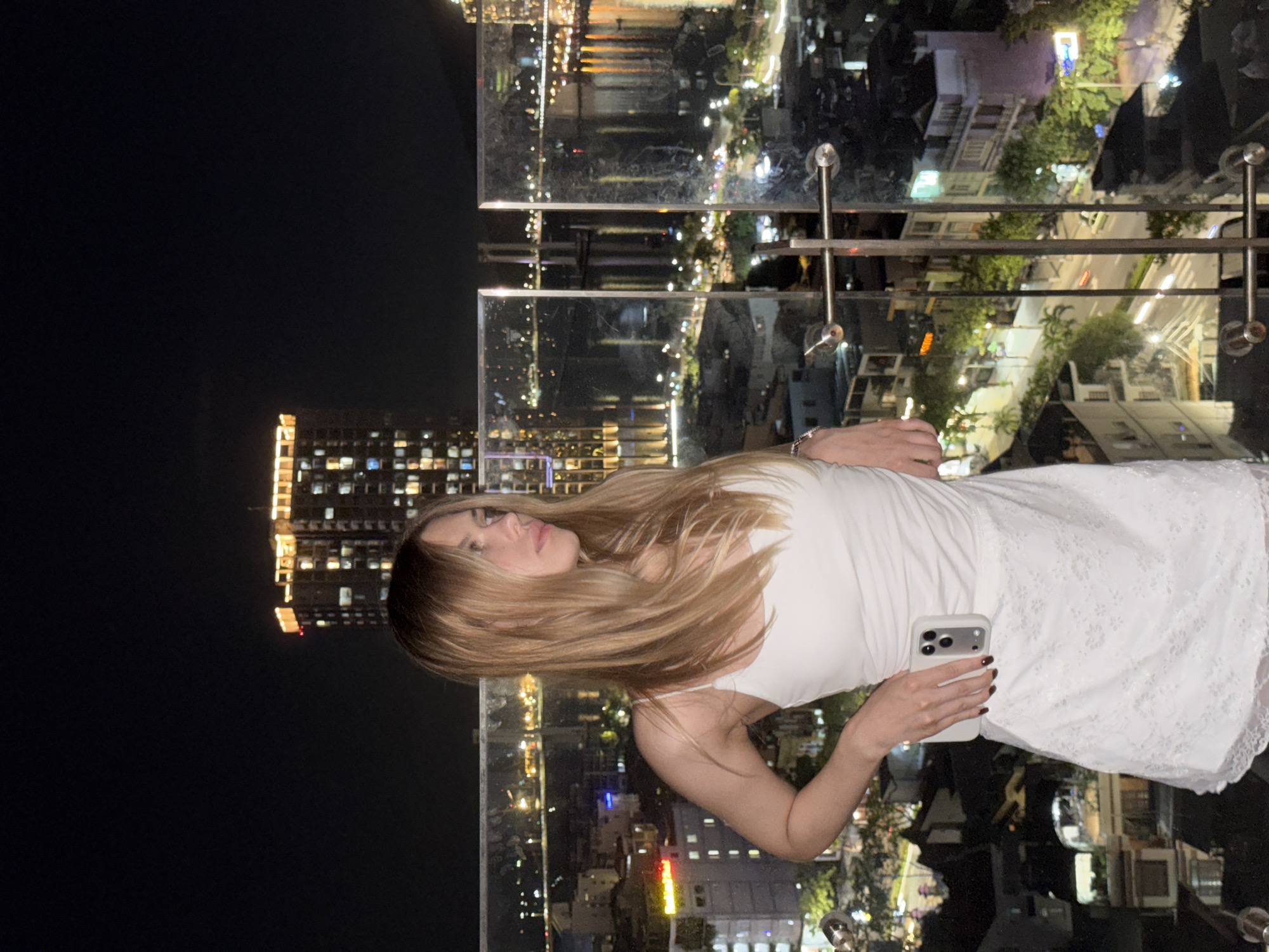 A young woman with blonde hair stands on a balcony at night overlooking the city of Đà Nẵng, Vietnam. She is wearing a white tank top and a white lace skirt. Her right hand is on her hip, and her left hand is holding a white smartphone. The city lights are visible in the background, with buildings, bridges, and a body of water. The sky is dark.