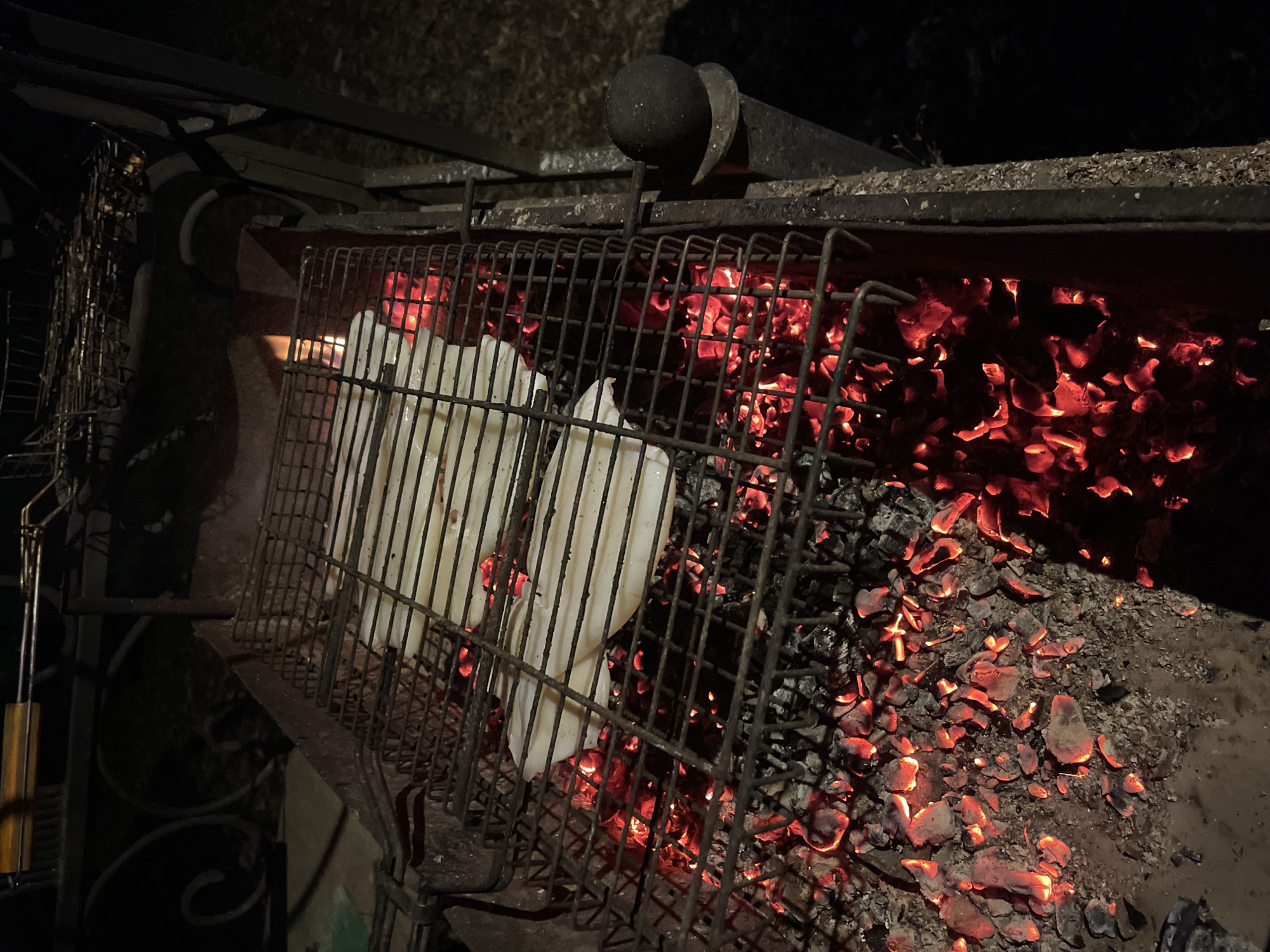 The image displays a nighttime outdoor grilling scene in Moscow, Russia. A metal barbecue grill occupies the center and right, filled with numerous glowing red and orange charcoal embers. These hot coals cast a warm light on the surrounding area and the grill structure.
Two rectangular metal grilling baskets are positioned over the embers. The basket on the left holds several light-colored, rectangular food items, which appear to be flat and uniform, possibly bread, cheese, or some form of dough, being cooked. The second basket, visible further to the right, is empty but also positioned above the heat.
The grill itself is constructed from dark metal, featuring a sturdy frame and various support bars. In the upper central portion of the image, a dark, spherical finial sits atop a vertical metal rod, likely an ornamental or functional part of the grill structure. To the far left, a portion of a wooden handle, possibly belonging to a grilling tool or part of the barbecue, is visible. The background is largely dark and indistinct, consistent with the nighttime setting. No discernible text is visible in the image.