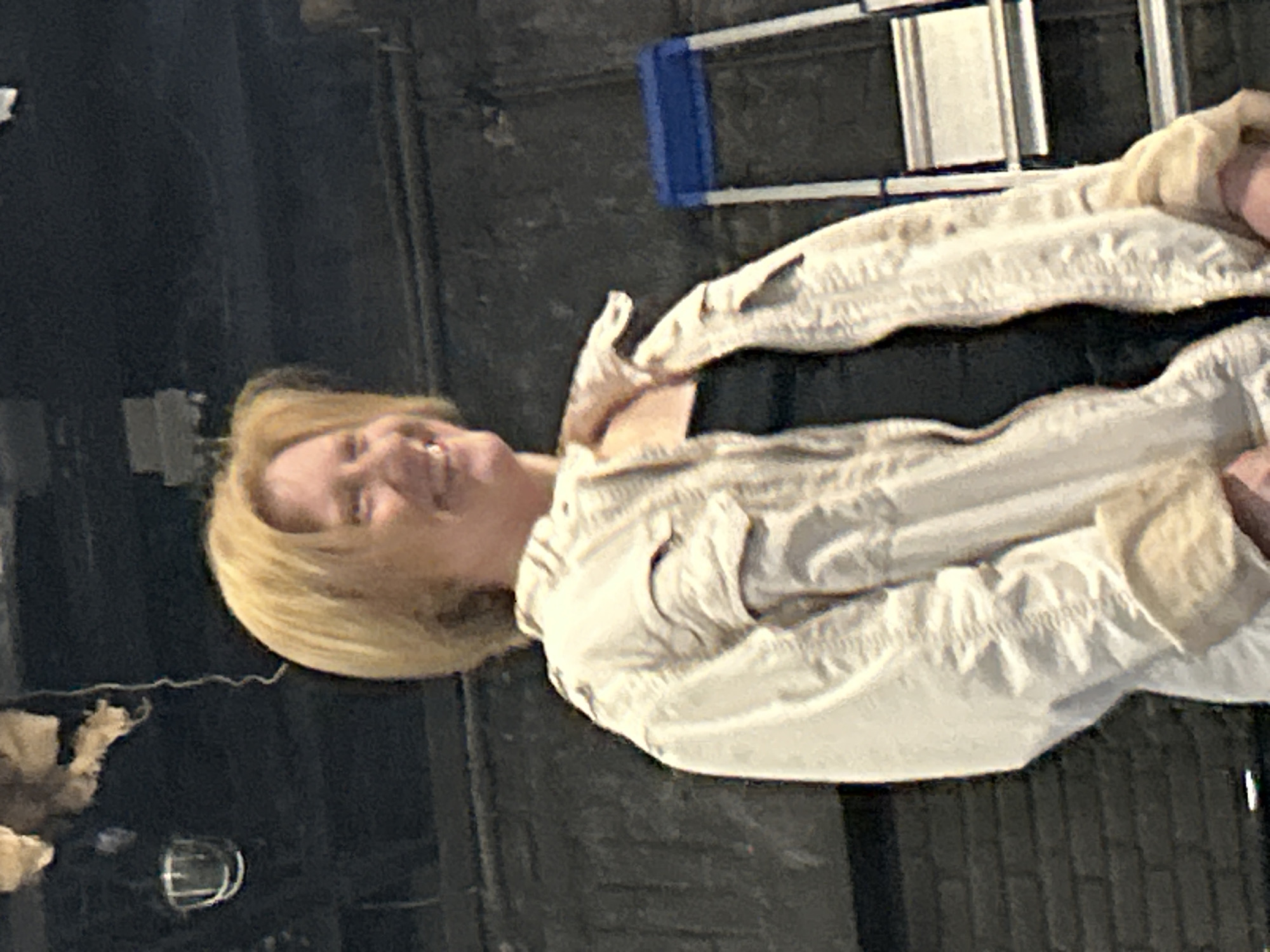 A woman with short blonde hair stands in front of a dark, textured wall. She is wearing a light-colored, possibly off-white, jacket over a black top. The jacket has a collared neckline and appears to be made of a fabric with visible stitching details, including flap pockets on the chest. Her right arm is bent and she is holding something that is not entirely visible. The woman is smiling and looking to her right. To the right of the woman, a blue and silver step ladder is visible, positioned against the wall. The background above the woman is dark and indistinct, with some hanging objects that appear to be some sort of theatrical or industrial lighting fixtures. The lighting in the scene is not uniform, with the woman being well-lit and the background receding into shadow. The overall impression is that the photo was taken in an indoor, possibly backstage or rehearsal space.