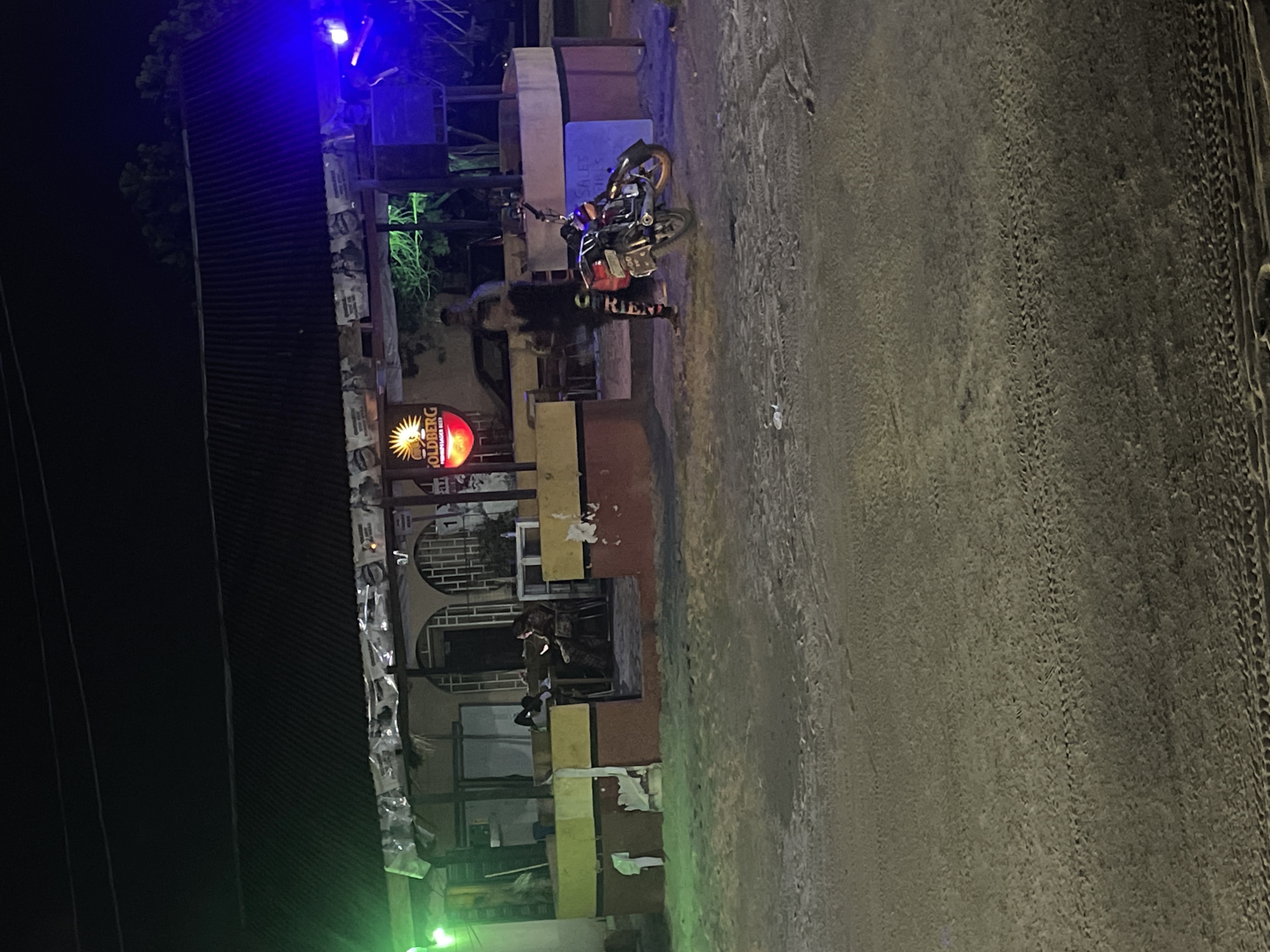 The image shows an urban street scene at night in Abraka, Nigeria. A brightly lit "GOLDBERG" sign is visible on a building. A person stands next to a parked motorcycle on the side of the road. The street appears paved with visible tire tracks. The buildings have multiple stories. There are electrical wires running along the side of the street. There are posters attached to the building in the upper-left. The sky appears dark. There are some lights coming from the building at the top of the picture. The image is tilted to the left.