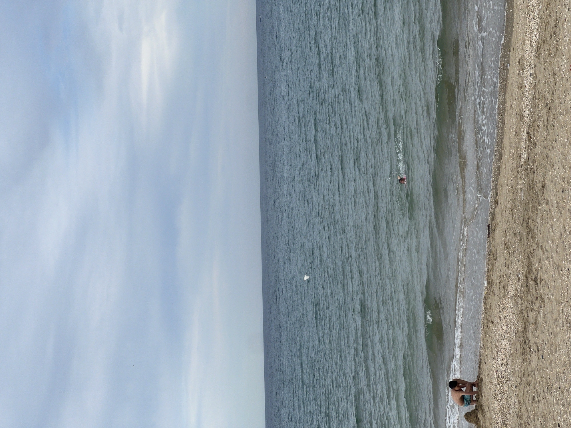 The image shows a beach in M'Diq, Morocco. The horizon separates a light blue sky with sparse clouds from a gray-blue sea. A person is swimming in the sea. Another person is crouched on the sandy beach near the water's edge. The beach is composed of sand and small shells. The image is oriented vertically.