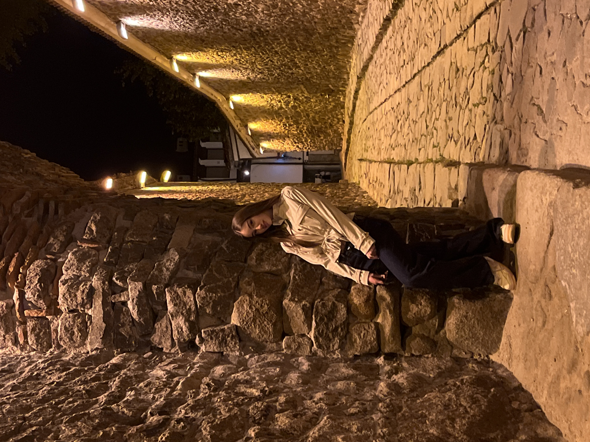 A woman lies on a stone wall in the city of Plovdiv, Bulgaria. She is wearing a light-colored jacket and dark pants. She is positioned along a stone wall next to ancient ruins lit by recessed lights. The walls are made of large, roughly cut stones, and a paved pathway lies in front of them. The background shows a dark night sky and a building with lights.
