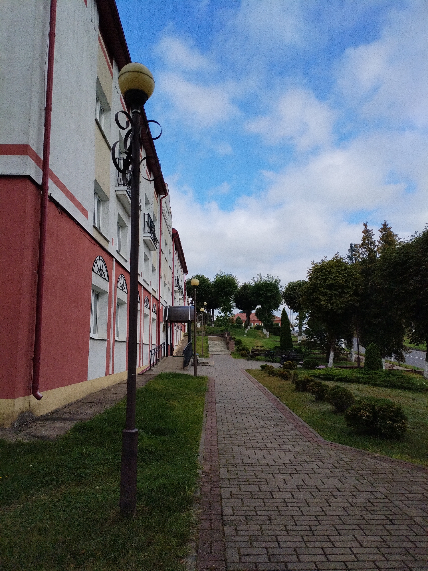 The image depicts a sidewalk in front of a red and white apartment building in Bialynichy, Belarus. The building has multiple windows and balconies, and a dark gutter runs down the side. A tall lamppost with a yellow spherical lamp stands on the grass next to the sidewalk. The sidewalk is paved with red and gray bricks and curves to the right, leading into a park area with trees, benches, and manicured bushes. The sky is blue with white clouds.