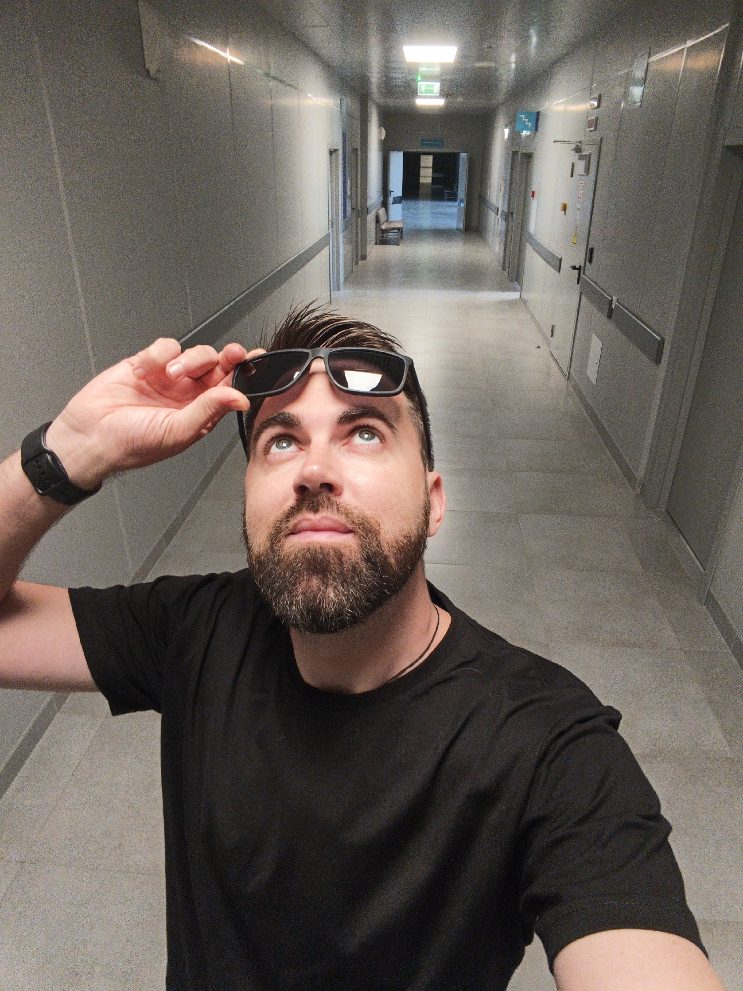 A man with a beard and dark hair is depicted in a close-up, taking a selfie or being photographed, as he looks upwards with a contemplative expression. He is wearing a black t-shirt and a black wristwatch on his left wrist, and holds up dark sunglasses with his right hand, resting them on his forehead.
The background reveals a long, straight, brightly lit hallway with light grey or beige paneled walls and a matching tiled floor. Fluorescent light fixtures are visible along the ceiling, providing illumination. Numerous doors line both sides of the corridor, and a handrail runs along the bottom of the wall panels. Towards the far end of the hallway, an open doorway leads into a less-lit area where a chair is partially visible.
Notable details include a green "exit" sign with a white running person pictogram visible on the ceiling in the distance. Another blue sign with a white upward-pointing symbol, possibly indicating stairs, is affixed to the right wall further down the hall. The setting appears institutional, sterile, and modern, reminiscent of a hospital, office building, or possibly a bunker. The lighting suggests an indoor environment. This scene is located in Балаклавский округ, Ukraine.