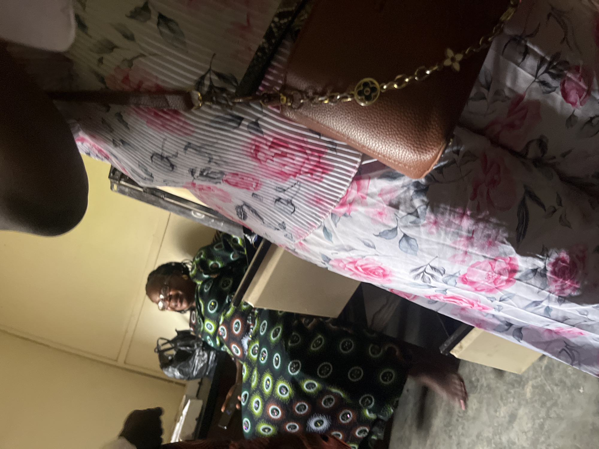 The image depicts an indoor scene with at least two women present. One woman, wearing glasses and a green patterned dress with circular designs, is seated and appears to be smiling. She is positioned in the background, to the left of the frame. In the foreground, a second woman, or part of her, is visible, wearing a light-colored dress with a floral pattern featuring pink roses. A brown purse with a gold chain is also visible in the foreground, hanging from her shoulder. The environment appears to be somewhat cluttered, with a black plastic bag on a surface behind the seated woman. The flooring is grey and appears to be concrete. The walls are a pale yellow. Without additional information, it is not possible to definitively place this scene in Jos, Nigeria, or any specific city.