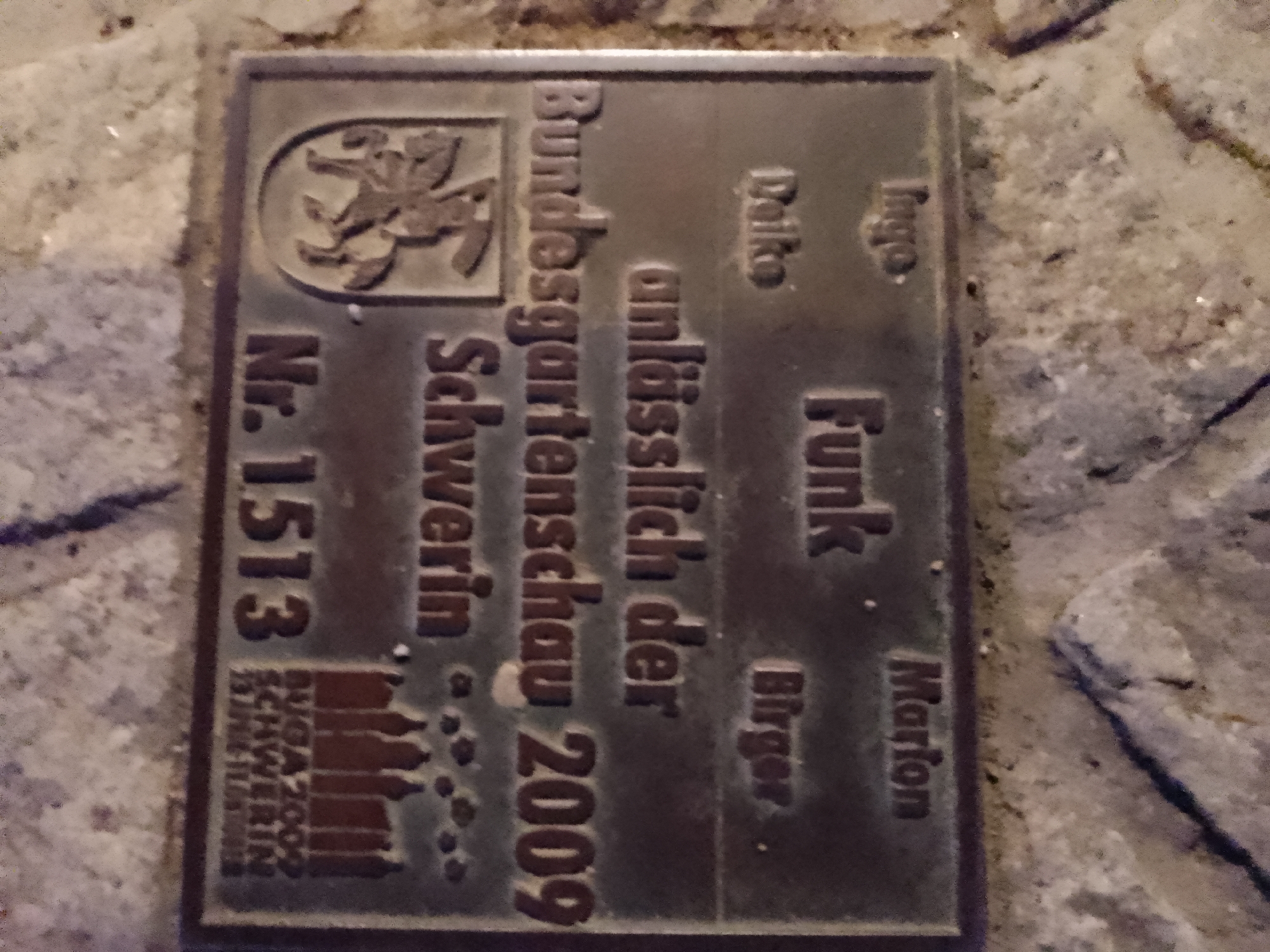 The image shows a metallic plaque attached to a stone surface. The plaque is inscribed with German text and a graphic. The text appears to be a dedication or memorial. Prominent elements include:
* **"Nr. 1513"**: This is a number, possibly an identifier or serial number, on the left side of the plaque.
* **"Bundesgartenschau Schwerin 2009"**: This indicates that the plaque is related to the Federal Garden Show held in Schwerin in 2009.
* **A graphic of a building facade**: Located near the bottom left of the plaque, this appears to be a stylized representation of buildings.
* **Names and a phrase**: The plaque also contains names like "Delko," "Ingo," "Funk," "Bürger," and "Marion." The phrase "unlässlich der" is also visible.
The stone surface appears to be part of a walkway or paved area. The lighting suggests it might be outdoors, possibly at night or in a shaded area. The plaque is weathered, indicating it has been exposed to the elements. The overall context suggests this is a marker or commemorative item related to a significant event in Schwerin.