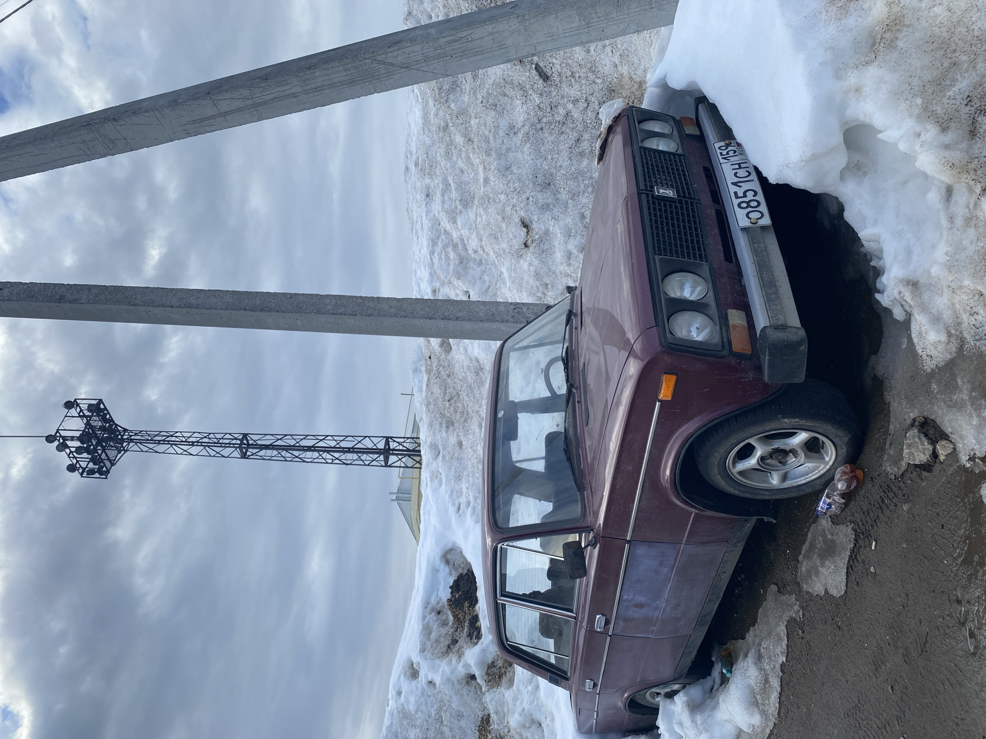 A maroon Lada 2106 sedan is positioned on a muddy, unpaved surface adjacent to large, dirty snow banks under an overcast sky in Chaikovsky, Russia. The vehicle displays visible grime and paint wear, notably a faded section on the driver's side front door. Its front license plate, "O851CH 159 RUS," is partially obscured by snow on the right side. Two rough, grey concrete utility poles lean diagonally, partially framing the scene. In the background, a tall, dark metal lattice tower, equipped with multiple spotlights at its apex, stands against the cloudy sky. A light-colored building with a sloped roof and multiple vents is partially visible behind the snow banks. On the ground near the front left tire, a clear plastic bottle containing a brown liquid and an empty clear plastic bottle are present, along with several discarded cigarette butts. No individuals or active interactions are depicted.
