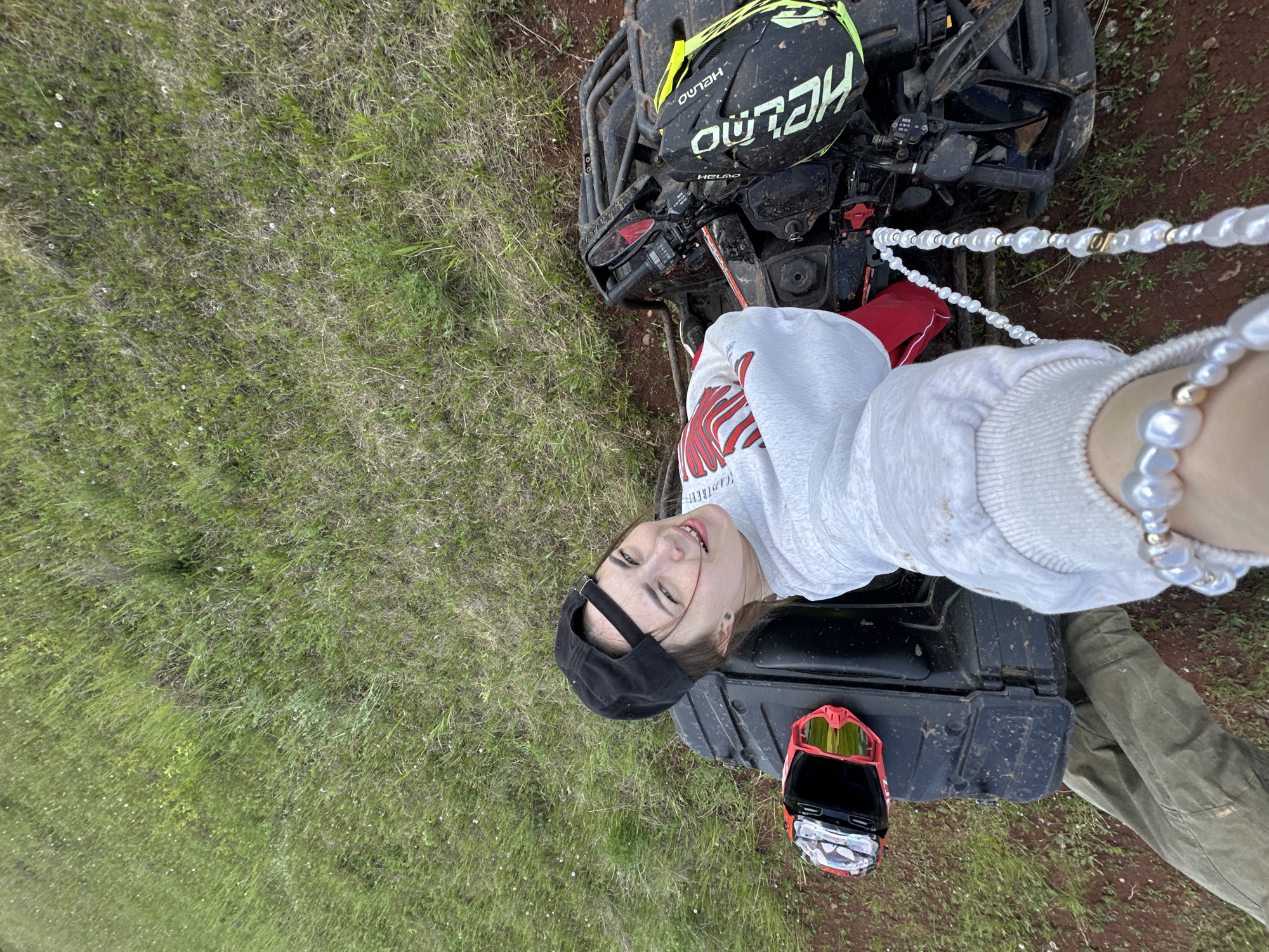 A young person is taking a selfie while positioned on a black all-terrain vehicle (ATV). The person is wearing a light-colored sweatshirt, a black cap, and a pearl necklace and bracelet. On the front of the ATV is a black and yellow helmet, and on the back is a red and black pair of goggles. The ATV is on dirt and grass. The location is likely Ilyinsky, Russia.