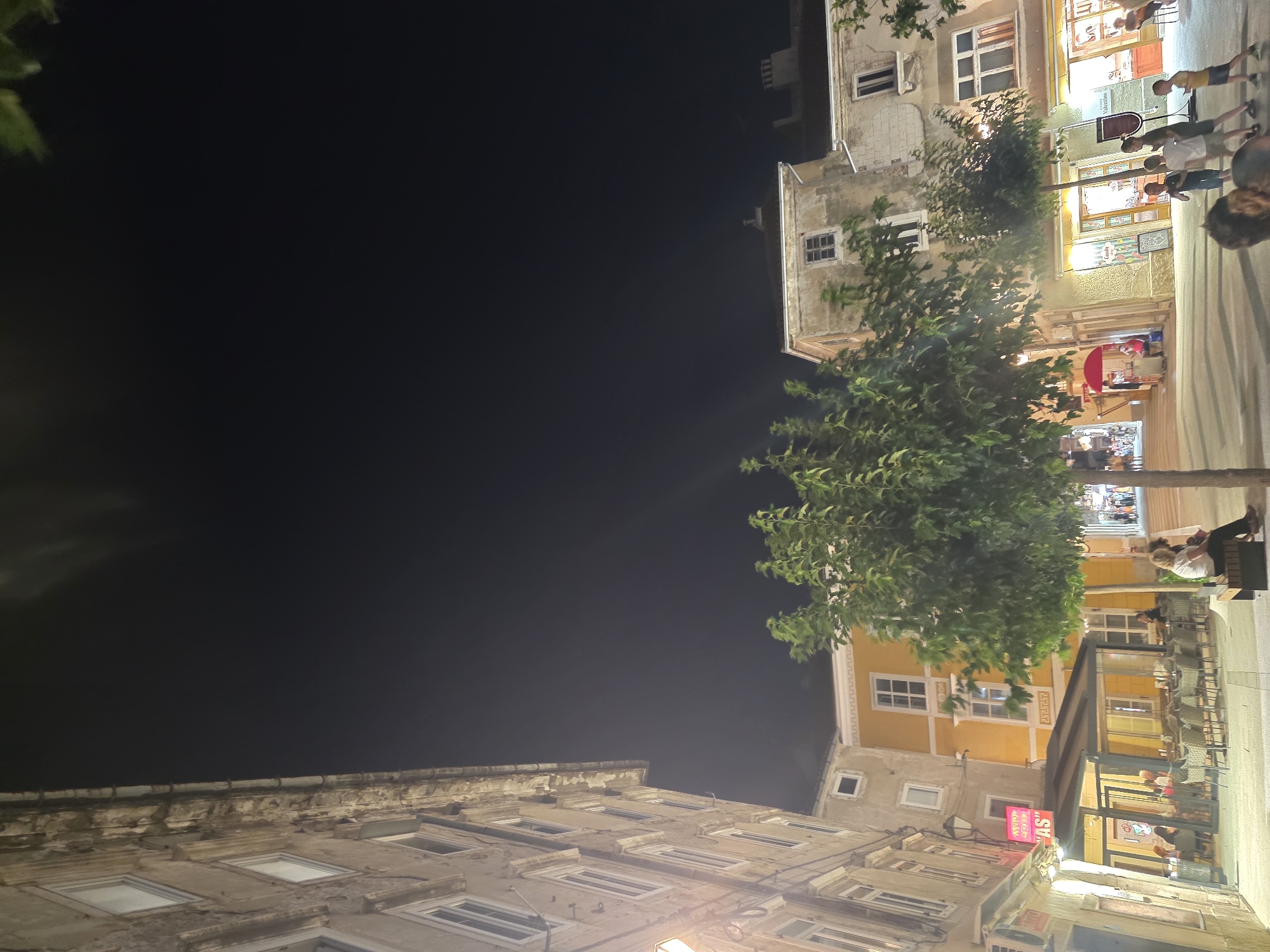 This is a low-angle, wide shot of a city square at night. On the left, a tall, weathered building with multiple windows and ornate trim lines the side of the square. Across from it, a bright yellow building with similar architectural details stands out. In the foreground, several trees with lush green foliage are scattered throughout the plaza, providing shade and visual interest.
Several people are visible in the scene. In the mid-ground, a group of children are playing in the square, while a few adults are seated at tables outside a cafe or restaurant. Another person is sitting on a bench near one of the trees. In the background, more people can be seen walking in the distance, suggesting a lively atmosphere in the square.
The lighting suggests it's evening, with streetlights and illuminated shop windows casting a warm glow on the scene. The overall impression is a pleasant, bustling urban environment.