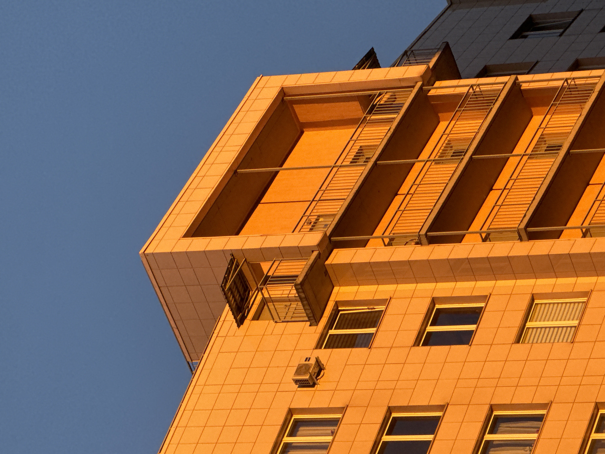The image shows a section of a modern building in an urban environment, possibly during late afternoon or early morning given the warm, golden light. The building's facade is tiled with a light-colored material that takes on an orange hue from the light source. Rows of windows are visible on the lower portion of the shot, some with blinds. Above the windows, there is a cantilevered structure with metal supports or railings that create diagonal patterns. A small ventilation unit is mounted on the wall between the window rows. The sky is clear and a deep blue color, contrasting with the warmth of the building. The location of the building within the city of Unknown, Unknown is not apparent from the image alone.