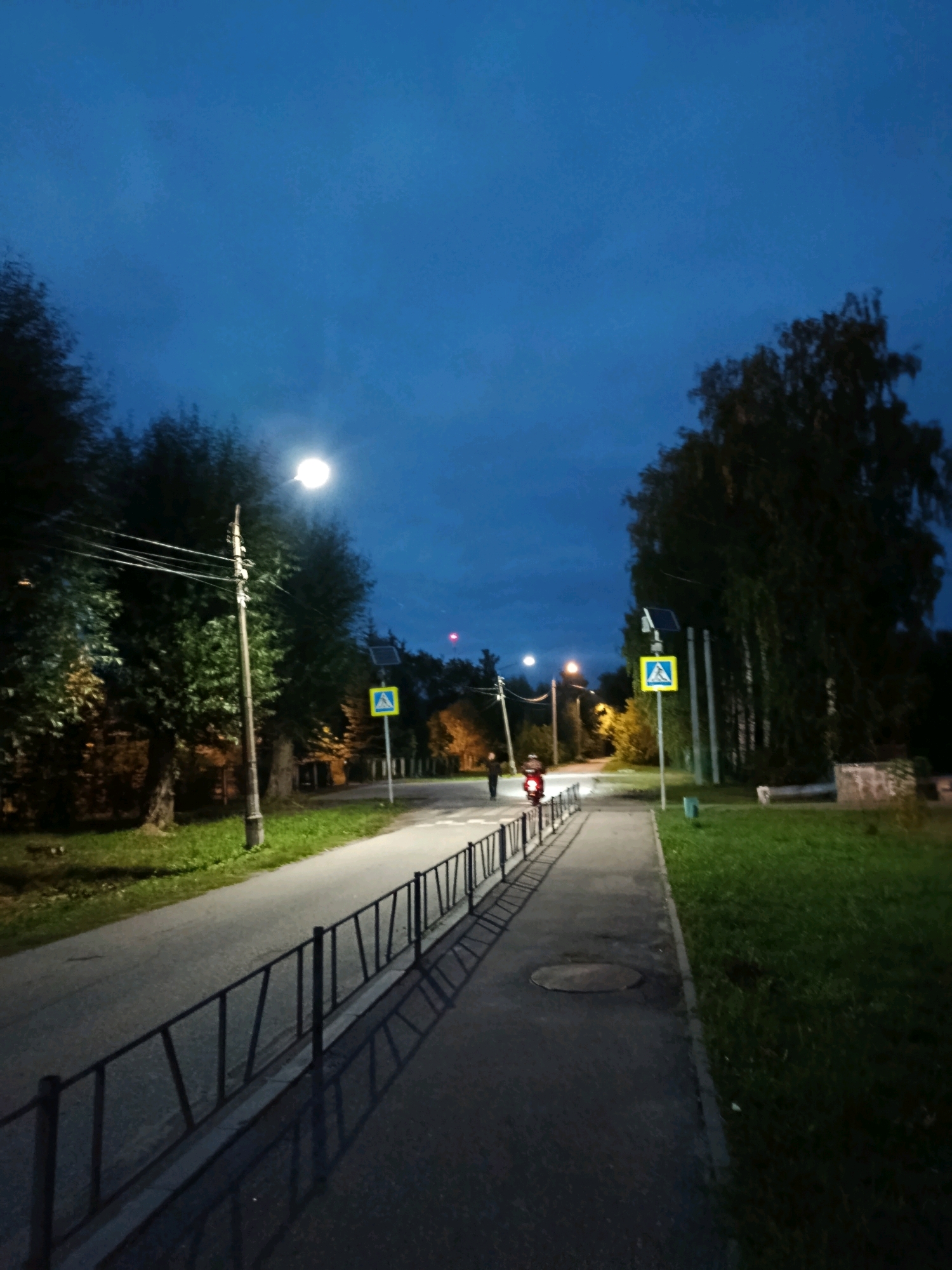In the city of Vereya, Russia, this image captures a twilight scene on a paved road with a sidewalk. A pedestrian crossing sign is visible on the right side of the road. Trees line the sides of the road, and streetlights illuminate the path. Two individuals are present; one on a red scooter rides down the road, and another person stands near the scooter. The sky is dark blue, indicating it is dusk or early night. A black metal fence runs along the edge of the sidewalk, casting long shadows on the pavement. The overall atmosphere is calm and quiet.