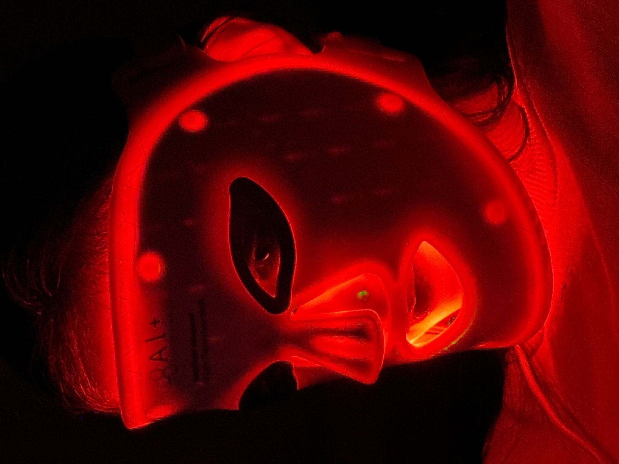 An upward-facing, close-up shot depicts a person undergoing a red light therapy treatment in a dark, intimate indoor setting. The central subject is a translucent red light therapy mask, glowing intensely, which covers the majority of the person's face. The mask features cutouts for the eyes, nose, and mouth. Small, circular light emitters are visible across its surface, contributing to the bright red illumination.
Faintly visible on the left side of the mask, the text "RAL+" is inscribed. Near the nose cutout, a small, distinct green light can be seen, possibly an indicator or status light. Dark hair is visible around the edges of the mask, particularly on the left side, confirming a person is wearing it. To the right of the mask, some reddish fabric, possibly bedding or clothing, is visible against the otherwise dark background. The scene is dominated by the strong red light emanating from the mask, suggesting the image was taken in low light conditions or at night, enhancing the glow of the device. No specific location cues are visible within the image itself.