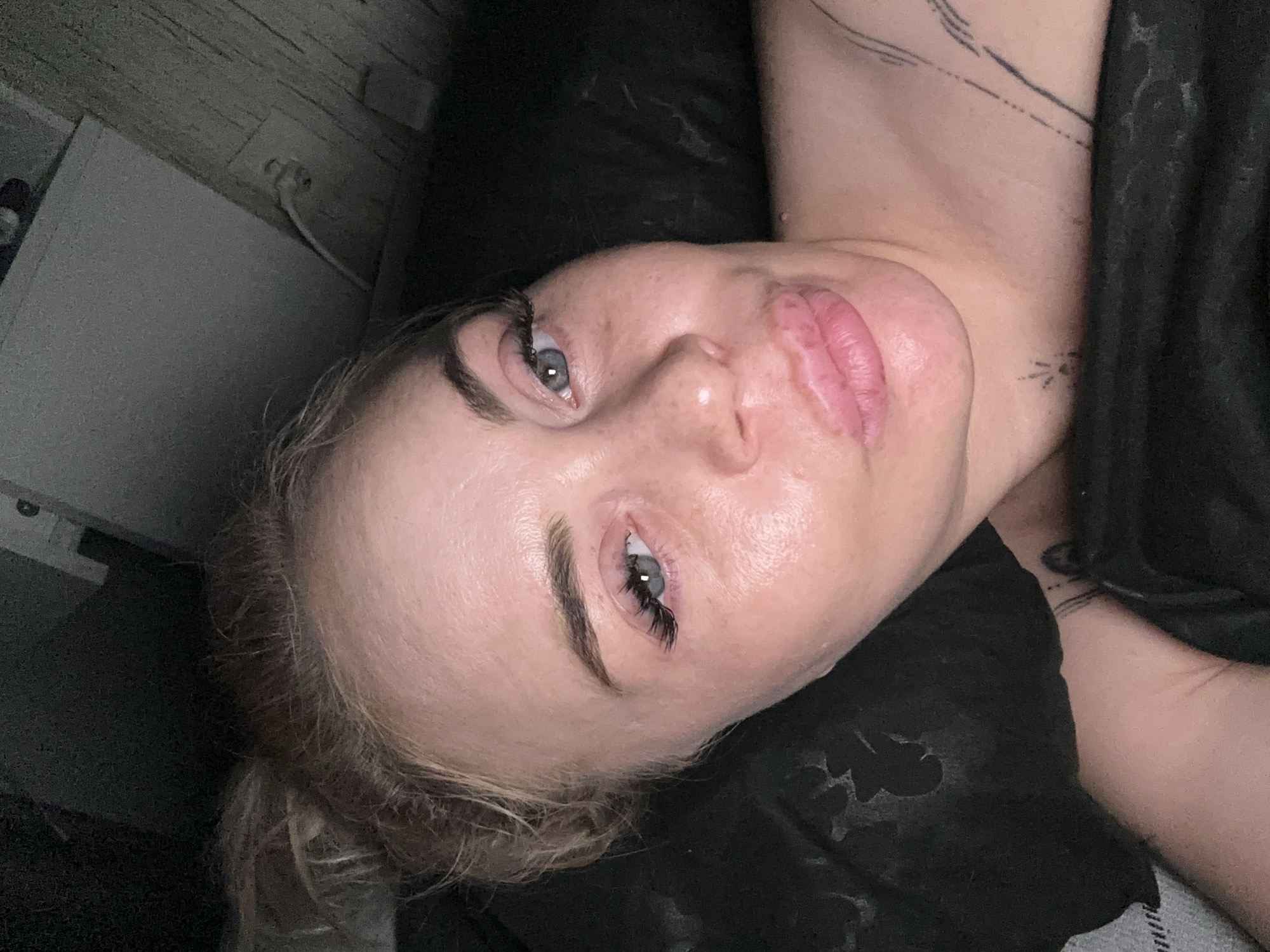A fair-skinned adult female is depicted from the upper chest upward, taking a selfie in a dimly lit indoor setting. Her blonde hair is gathered into a messy bun on top of her head. She has blue-grey eyes with prominent dark eyelashes, and well-defined dark eyebrows. Her lips are full and pursed in a "kiss" expression. Visible on her upper torso are multiple fine-line tattoos, including parallel lines on her left shoulder and a small starburst design centrally on her chest/neck. She is draped in dark fabric. The background to the left is dark and indistinct. To the upper right, a light grey rectangular object is visible above a textured wall, which features an electrical outlet with a white cord plugged in. The location is in the city of Unknown, Unknown.
