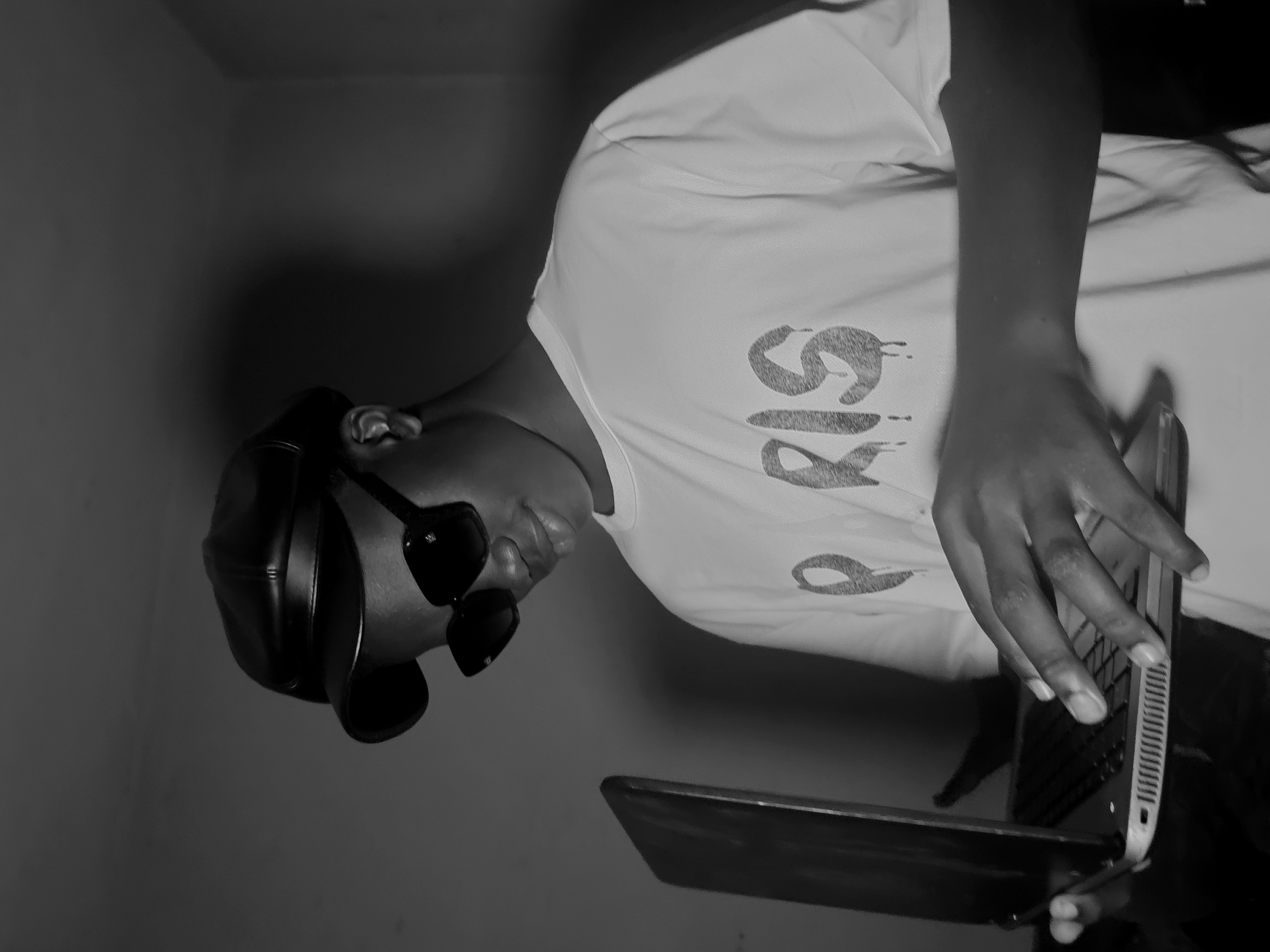 A person is shown in a black and white photograph, holding a laptop. They are wearing sunglasses and a hat. The background shows a plain wall. It is not possible to ascertain a specific location in Nkwelle, Nigeria, or confirm that the image was taken there.