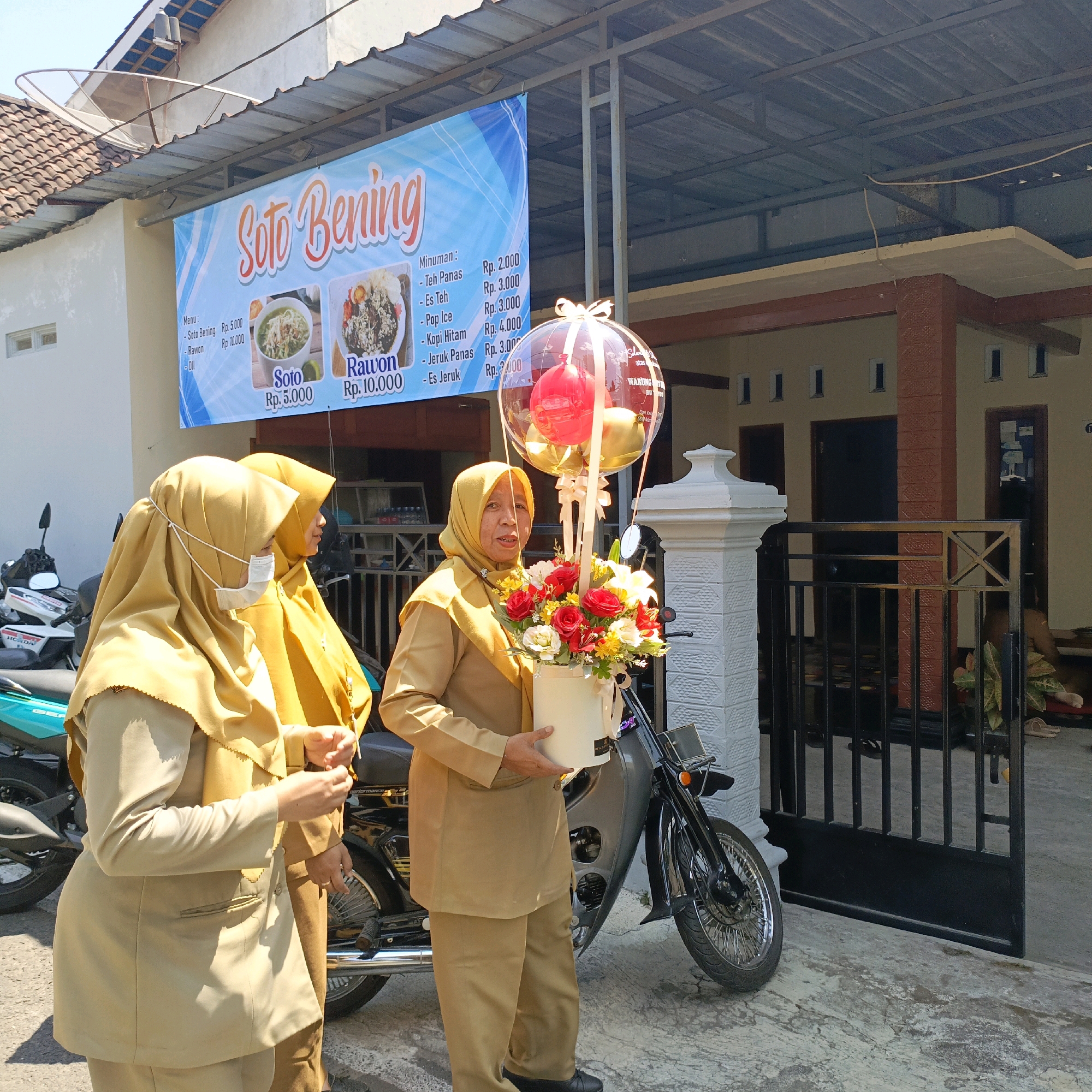 A group of three women, all wearing tan uniforms and yellow hijabs, stand in front of a building with a banner that reads "Soto Bening" and lists menu items and prices. The woman in the foreground on the right holds a white bucket filled with red and yellow flowers, and a clear balloon filled with smaller red and gold balloons. She is looking towards the camera and speaking. The woman behind her to the left is wearing a white face mask and also looking towards the camera. The third woman, partially obscured, stands to the left of the woman with the flowers. Two motorcycles are parked behind the women. The building appears to be a shop or restaurant. The setting is outdoors, likely in a residential or commercial area of Salatiga, Indonesia, given the banner's text and the style of architecture.