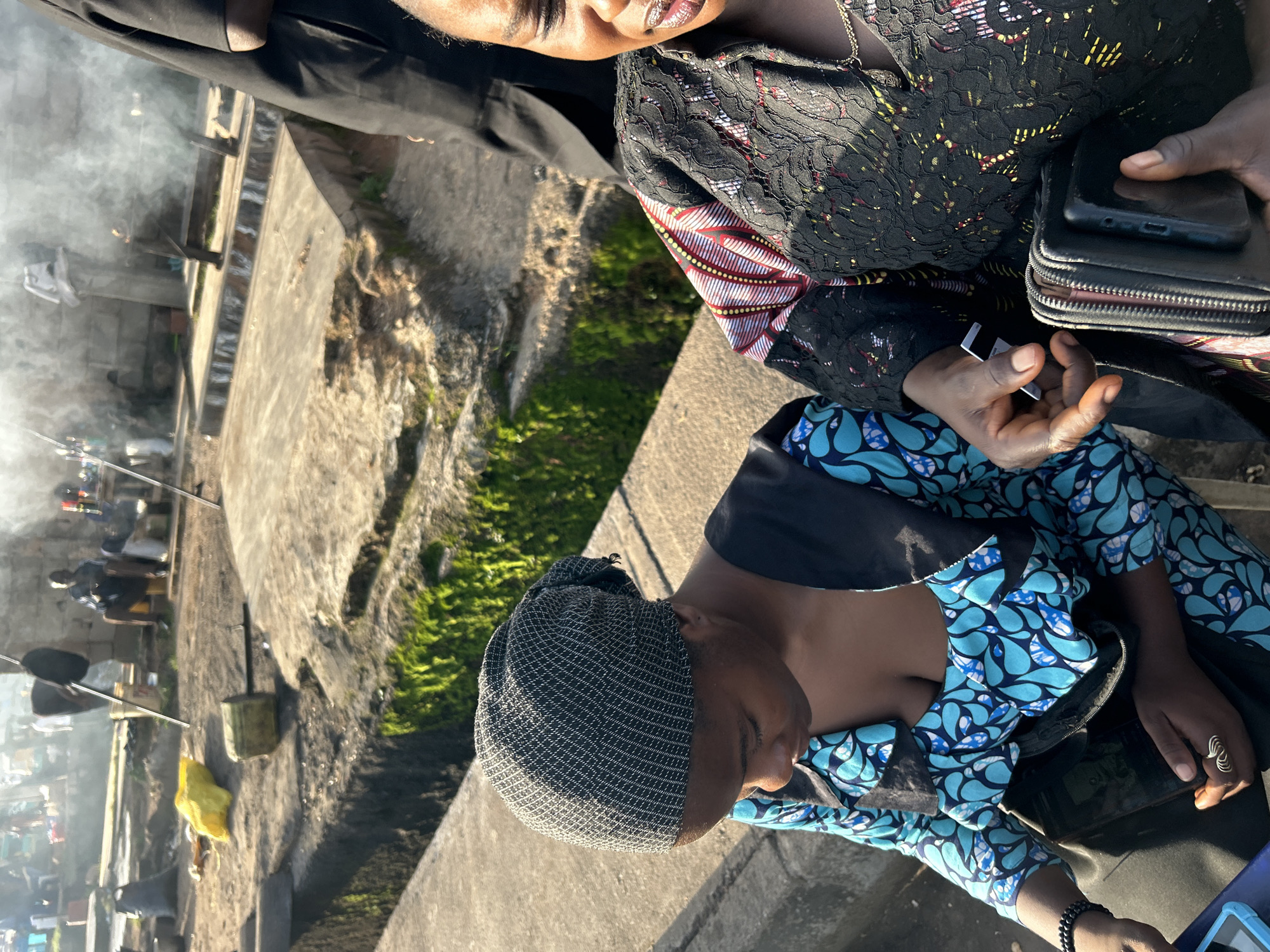 In Jos, Nigeria, two women are seated on a concrete ledge, with a backdrop of an outdoor area featuring smoke emanating from what appears to be a cooking or burning activity. The woman on the left wears a patterned blue and black dress and a dark head covering. She is looking down at a dark bag resting on her lap and is holding a cell phone. The woman on the right is wearing a black lace top with colorful, patterned sleeves. Her mouth is open, and she is holding a black wallet in her hands. Behind them, the environment is rough and unpaved, with various objects scattered around, including bags, containers, and concrete structures, suggesting a functional or possibly informal area of the city.