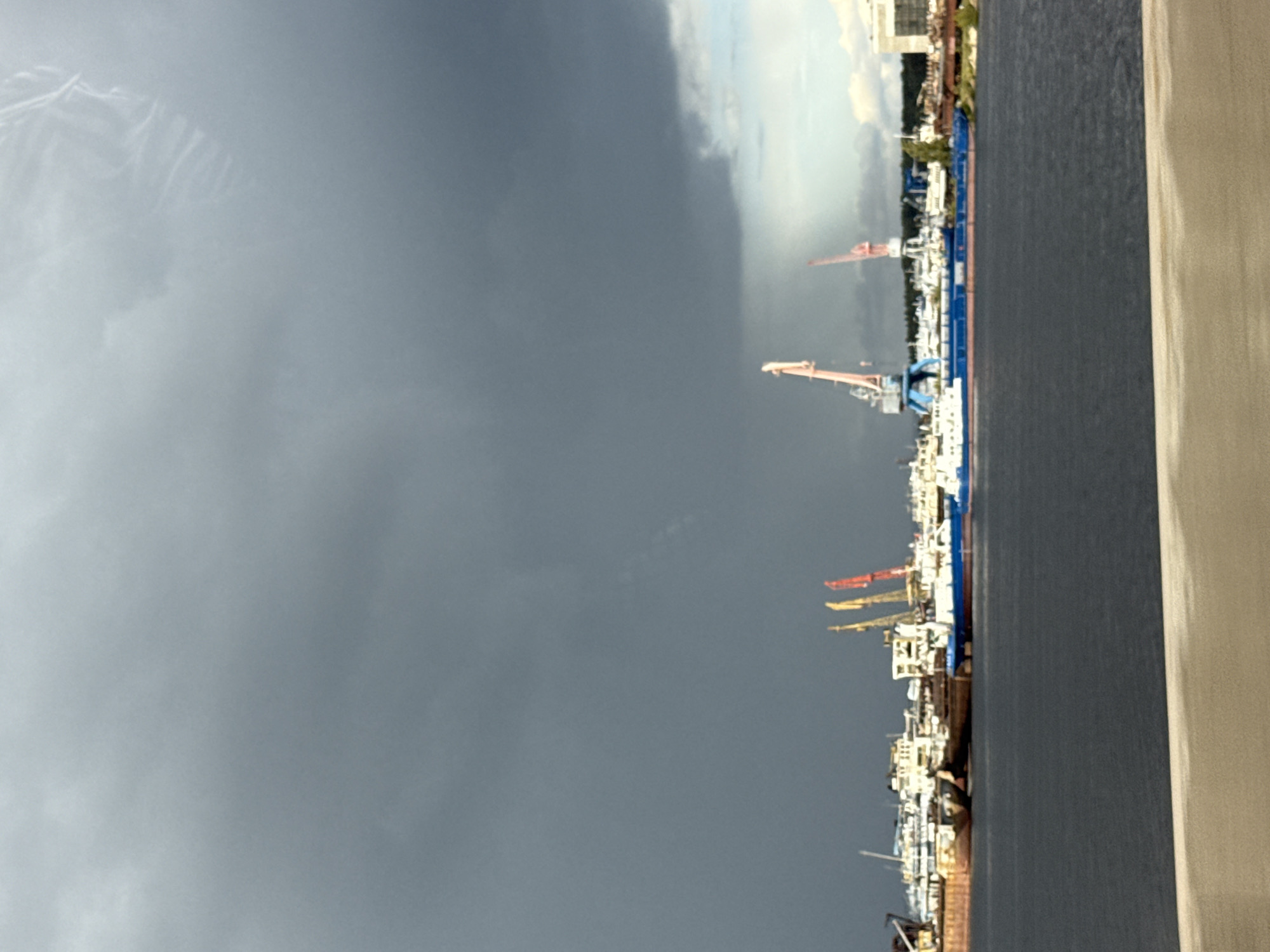 A cloudy sky looms over a harbor in Chaikovsky, Russia. Several ships and boats are docked, with some appearing to be cargo vessels. Cranes, one large blue and pink one and two smaller pink ones, stand on the docks, ready for use. The water in the harbor is dark and calm. In the distance, buildings and trees are visible, partially obscured by the dark clouds. The scene suggests a working port, possibly preparing for or experiencing inclement weather due to the heavy cloud cover.