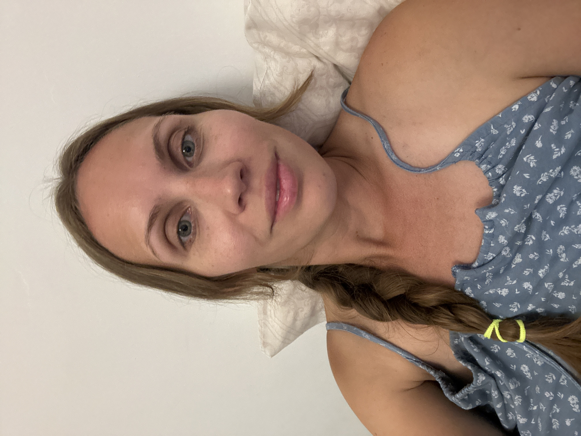 A woman lies on a pillow, facing the camera. She has light brown hair braided on one side, secured with a yellow band. She wears a blue floral patterned top with thin straps. The background consists of a white wall and a pillow with a subtle pattern. The image provides no information about the location of the subject in Fethiye, Turkey.