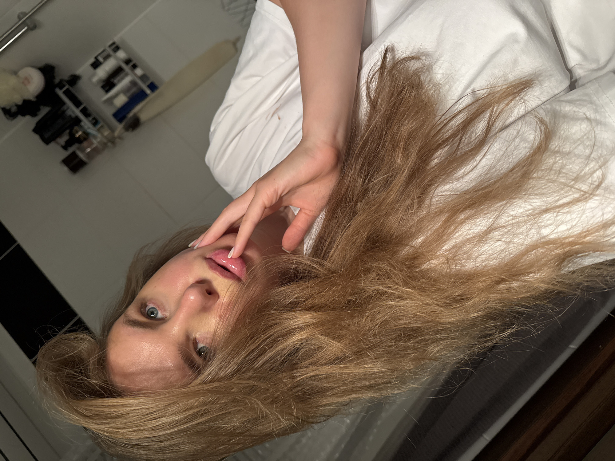 The image shows a close-up, high-angle shot of a young woman with long, wavy blonde hair. She is wearing a white t-shirt and her face is partially obscured by her hair. Her right hand is touching her lips, which appear to be glossed. Her eyes are blue and looking upwards. The background appears to be a bathroom with shelves holding various toiletries, including bottles and jars. The lighting is dim, with a strong light source coming from the left, casting shadows. The overall impression is that of a candid, intimate portrait. The location in the city of Vladimir, Russia is not discernible from the image.