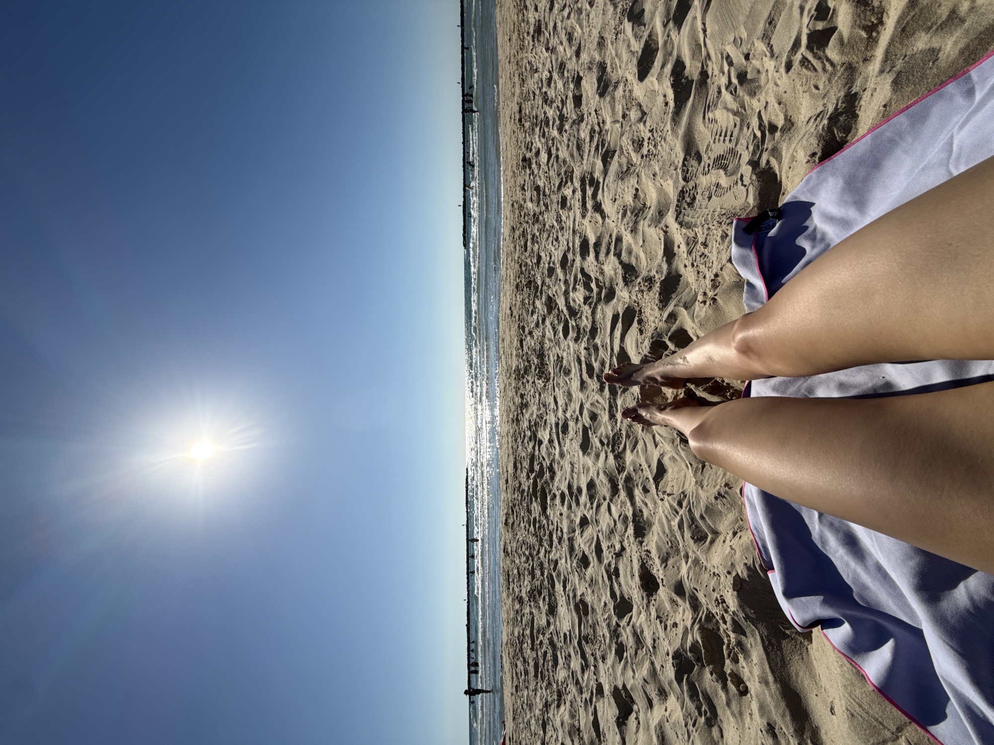 This is a high-angle, vertical shot of a beach scene on a sunny day. The image is divided into two halves. The left side shows a bright blue sky with the sun prominently featured, casting a lens flare and rays of light. The sun appears to be mid-morning or late afternoon. The right side of the image displays a sandy beach with footprints and ripples in the sand. A light purple beach towel with a pink trim lies on the sand, partially covering the legs of a person lying down. The person's tanned legs and feet are visible, suggesting they are enjoying a day at the beach.
In the distance, where the sky meets the sea, a faint line of people can be seen along a pier or breakwater. The water is calm and reflects the sunlight, creating a shimmering effect. The overall mood of the image is one of relaxation and enjoyment of a summer day. The setting is clearly a beach, likely a popular one given the presence of other people in the distance. No visible text is present in the image.