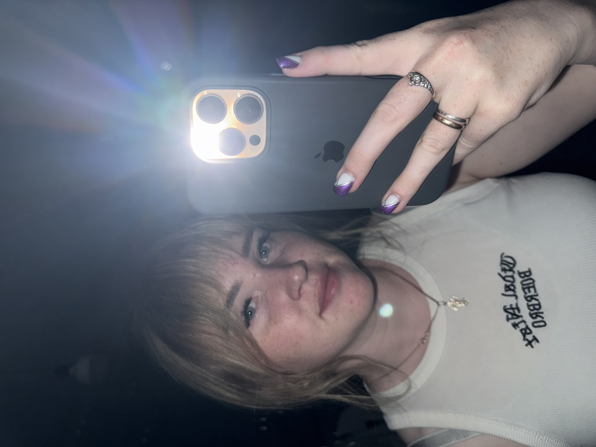 A selfie taken by a woman using a smartphone with a bright flash. She is wearing a white top with Cyrillic lettering on it and a necklace with a pendant. She has blonde hair with bangs and freckles on her face. The nails on her hand are decorated with purple and white designs. There are multiple rings on her fingers. The location cannot be determined from this image.