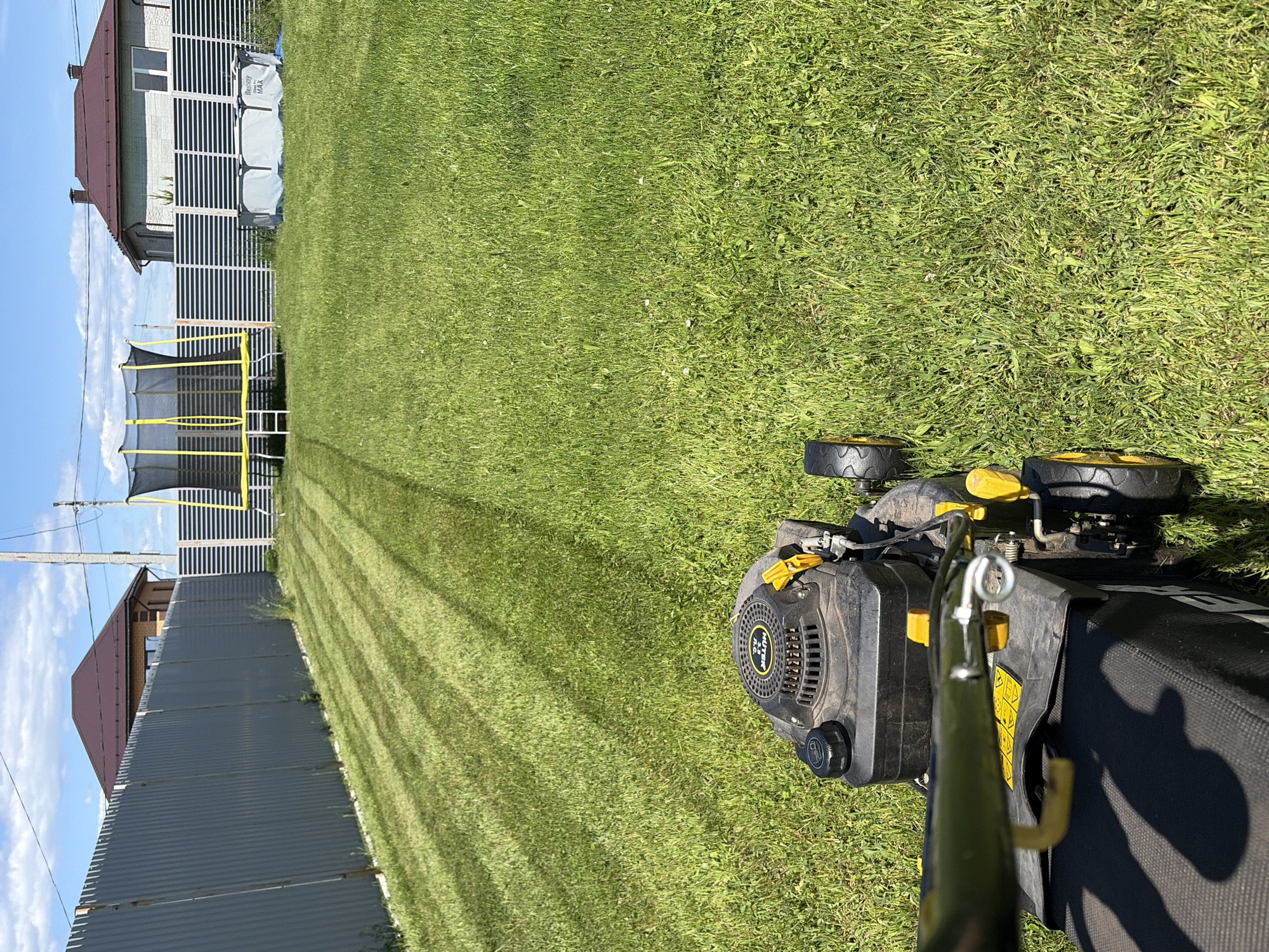 The scene depicts a first-person perspective of lawn mowing in a residential backyard in Chaikovsky, Russia, under clear blue skies with scattered white clouds. A black and yellow HUTER R.C. push lawnmower is partially visible in the foreground, with its engine cover, fuel cap, and part of the handle and grass catcher bag prominent. The right front and rear wheels are visible. The grass directly in front and to the right of the mower is unkempt, long, and green, interspersed with weeds and small white flowers.
In the midground, the lawn shows distinct stripes on the left, indicating areas that have been recently mowed. This mowed section is shorter and darker green, contrasting sharply with the unmowed grass that covers the right side of the yard and extends into the foreground.
The left background features a tall, dark grey corrugated metal fence running horizontally across the property boundary. Behind this fence, the upper portion of a house with a brown hip roof is visible. Further into the background, a utility pole stands against the sky.
The right background reveals a different fence, constructed from light-colored horizontal slats. Behind this fence, a large trampoline with a black net enclosure and yellow support poles is situated on the left. To its right, a light grey above-ground swimming pool, labeled "Bestway MAX," is present. Behind this fence and objects, another house with a brown hip roof and light-colored siding is visible, featuring white-framed windows. The overall environment suggests a sunny, fair-weather day suitable for outdoor activities.