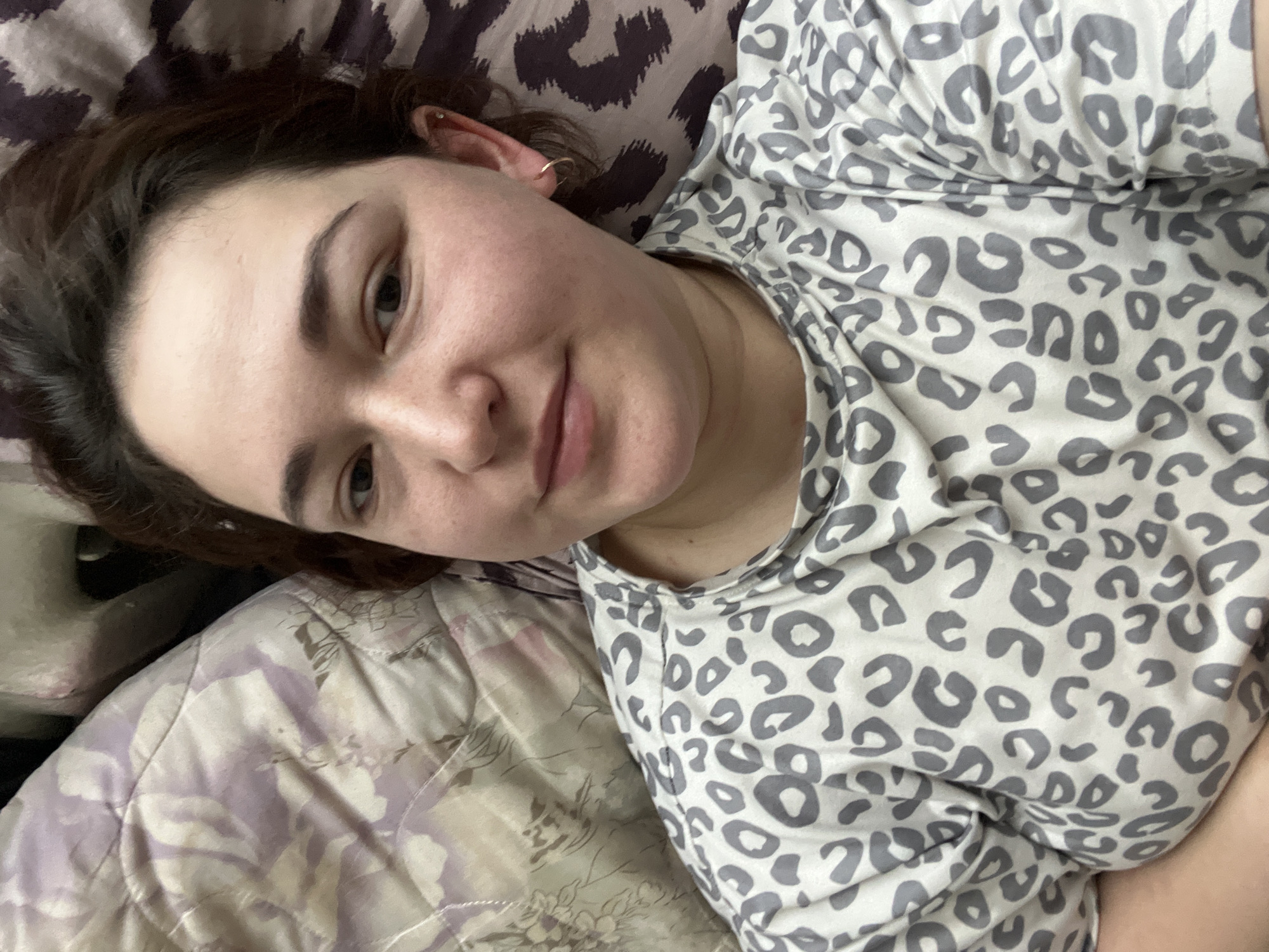 The image shows a close-up, overhead shot of a young woman lying down in bed. She has dark brown hair, fair skin with some freckles, and is wearing a white t-shirt with a grey leopard print. Her head is resting on a pillow with a floral pattern and another pillow with a black and white pattern is visible behind her. She is looking directly at the camera with a neutral expression. There is no indication of the location within the city of Chelyabinsk, Russia.