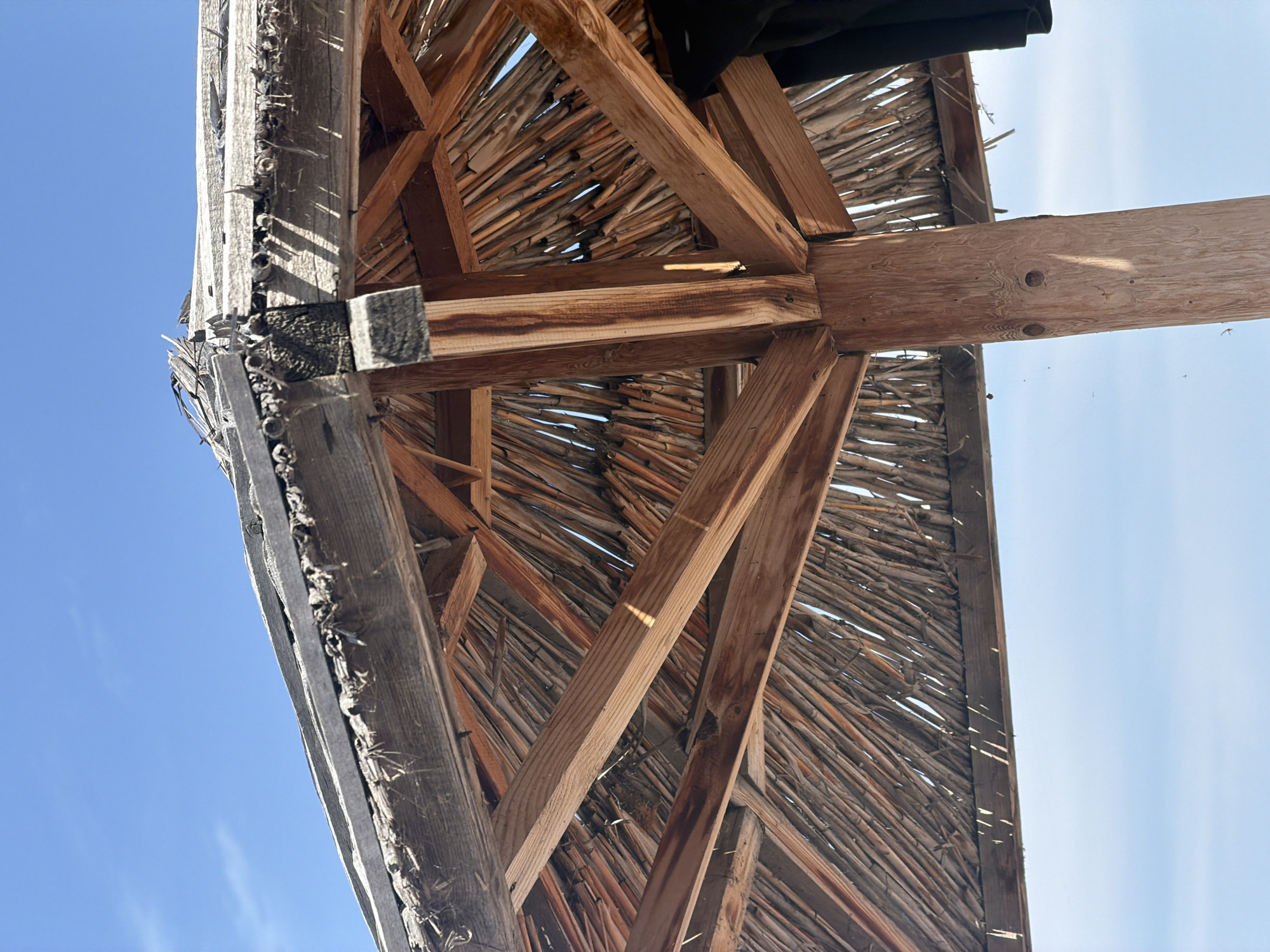 The image shows the underside of a thatched roof umbrella against a clear blue sky. The umbrella is constructed with wooden beams and posts, with dried reeds or straw forming the thatch. The wood appears weathered and aged, with some sections showing wear and tear. A dark piece of fabric, possibly clothing, is visible hanging on the right side of the image. The scene is likely outdoors, suggesting a sunny day. The location is specified as Choktal, Kyrgyzstan.