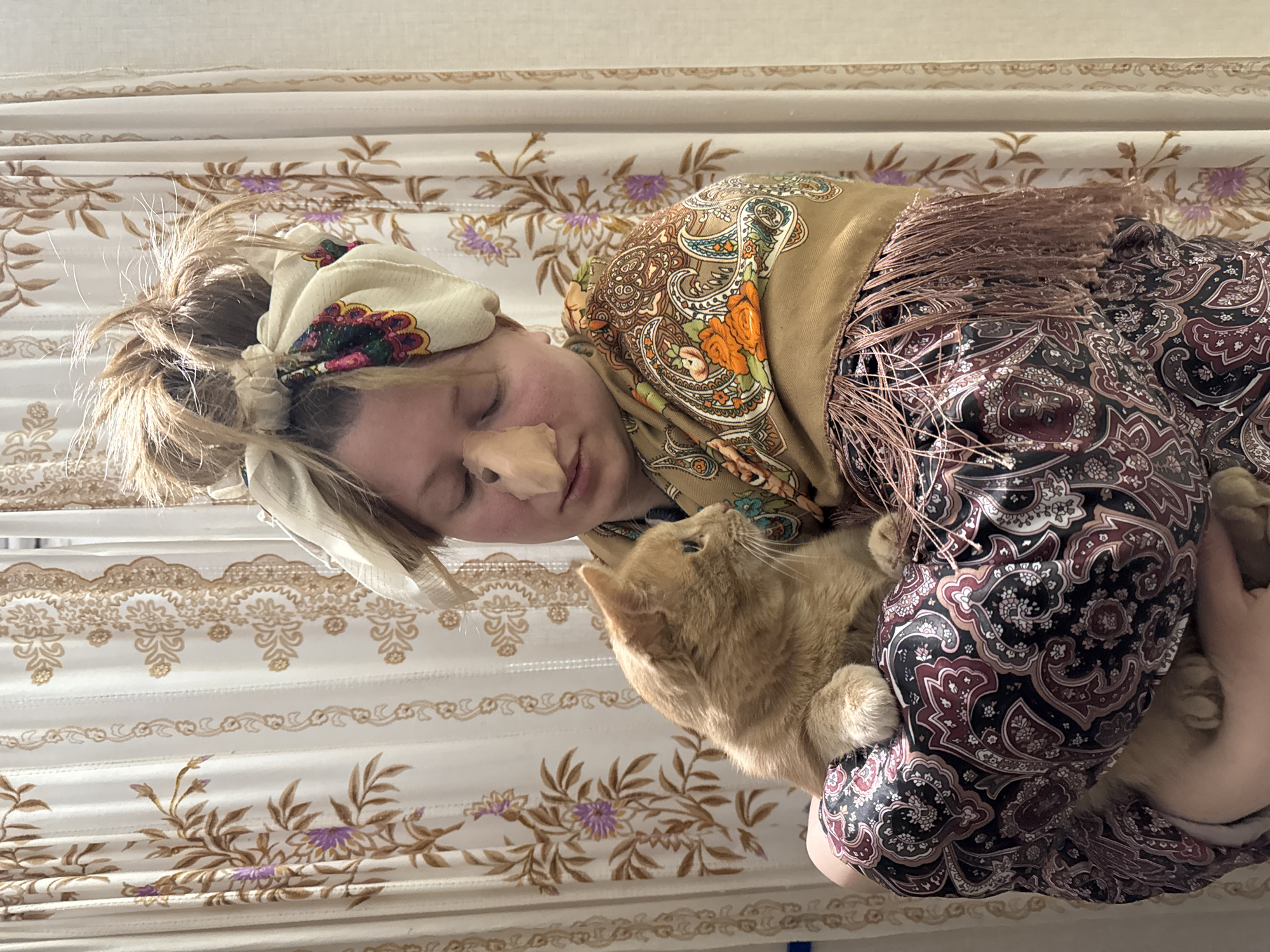 This indoor image from Вассята, Russia, captures a serene moment between a person and an orange cat. A person, appearing to be a woman, is lying down with her eyes closed, suggesting rest or sleep. A light-colored patch or material is visible on her nose.
She is dressed in a style reminiscent of traditional attire. Her head is wrapped in a cream-colored headscarf featuring a vibrant floral and paisley pattern, allowing some light brown hair to be seen. Over this, a larger, golden-brown shawl with an elaborate paisley and floral design in warm tones of orange, teal, and brown is draped, characterized by its long, fringe-edged border. She wears a garment with a dark background, intricately patterned with paisley and floral motifs in shades of maroon, black, and cream.
An fluffy orange tabby cat is curled up next to her, seemingly resting or being cuddled. The cat's front paws are visible, and its head is turned upwards and slightly away, observing something out of frame, yet appears calm.
The setting consists of light-colored fabric, possibly bedding or curtains, adorned with delicate floral patterns in muted gold, brown, and subtle hints of purple. The lighting suggests it is daytime, casting a gentle, natural glow on the scene. The overall impression is one of peacefulness, comfort, and affectionate companionship. There is no visible text in the image.
