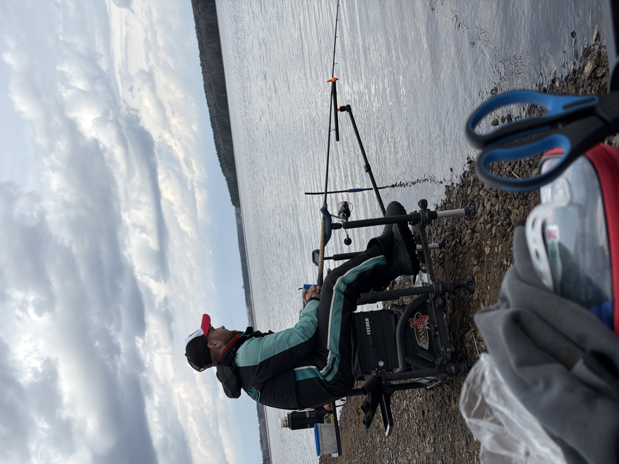 A man in a black and blue jacket, black pants, and black boots sits in a black fishing chair with a red and white hat, looking towards the cloudy sky. A fishing rod is set up on a stand to the right. To the right of the chair is a pair of blue handled scissors and a red bag. The ground is rocky and leads down to a body of water. A line of trees is visible across the water. The photo was taken in the city of Chaikovsky, Russia.