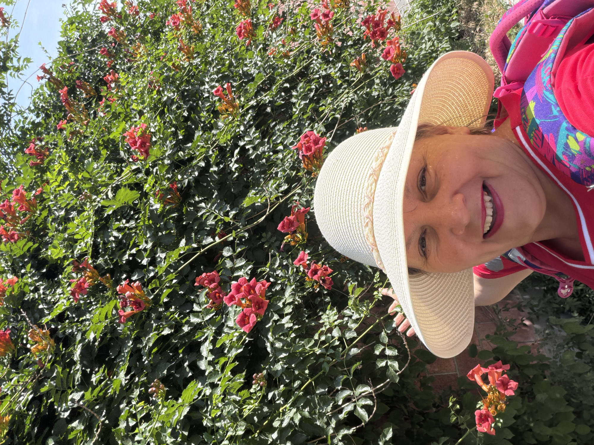 A woman wearing a straw hat and a bright red shirt smiles at the camera. She is standing in front of a wall covered in lush green foliage and vibrant red trumpet-shaped flowers. A colorful backpack is visible on her shoulders. The image appears to be taken outdoors on a sunny day.
