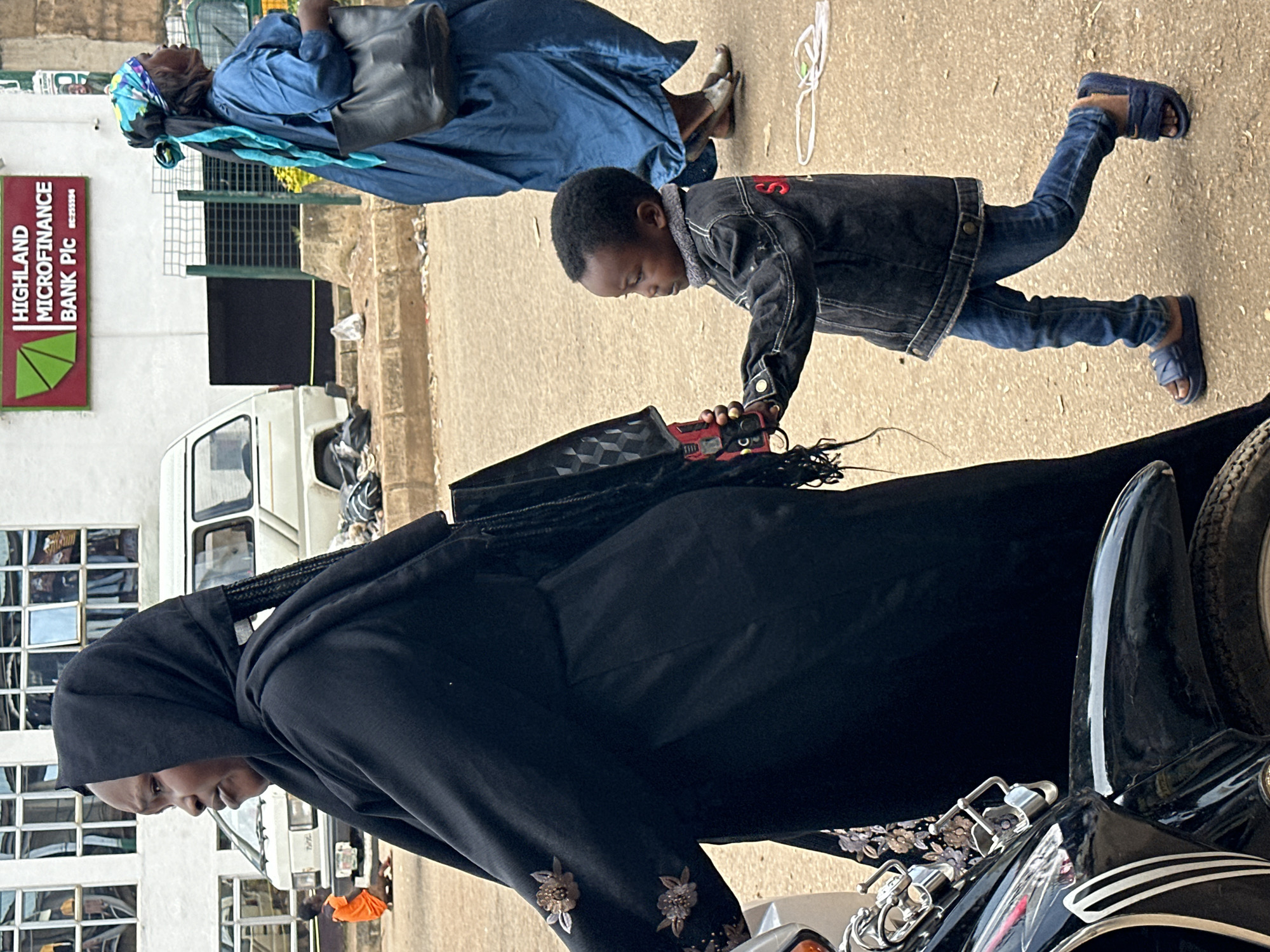 The image captures a street scene in Bukuru, Nigeria, focusing on three individuals and a parked motorcycle. In the foreground, a young child, appearing to be a boy with dark skin and short, dark hair, is walking to the right, holding the hand of an adult. The child is wearing a dark denim jacket over a light-colored shirt, blue jeans, and dark blue slide sandals. To the left of the child, an adult, a Black woman, is walking away from the camera and towards the left. She is wearing a black headscarf and a flowing black abaya with decorative floral appliques on the sleeves. A black bag with a patterned design is slung over her right shoulder, and part of a motorcycle is visible in the lower left corner, suggesting the adult may be associated with it. In the background, another Black woman is walking to the right, wearing a blue dress and a colorful headscarf, carrying a black handbag. Behind the individuals, a building with multiple windows is visible, and a white van is parked. The ground is paved with asphalt or dirt. The scene appears to be during daylight.