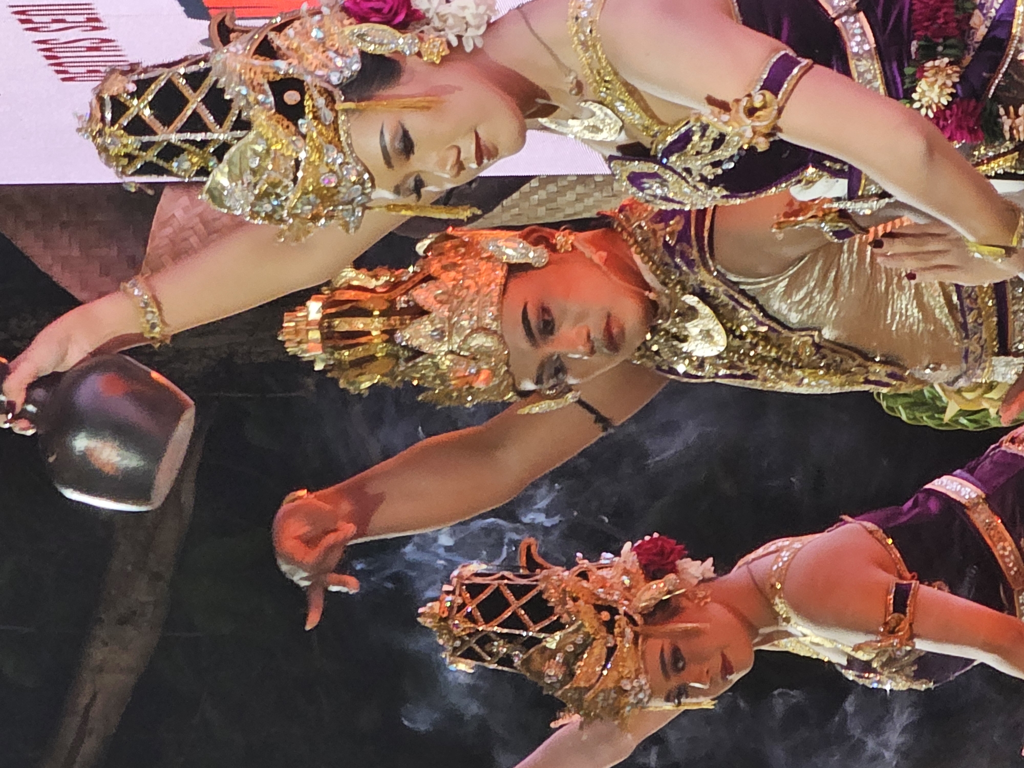 Three female dancers perform a traditional Balinese dance in Denpasar, Indonesia. The dancers are adorned with ornate golden headdresses, intricate jewelry, and purple and gold costumes. One dancer holds a dark ceramic pot. Smoke swirls around them. The background is dark.