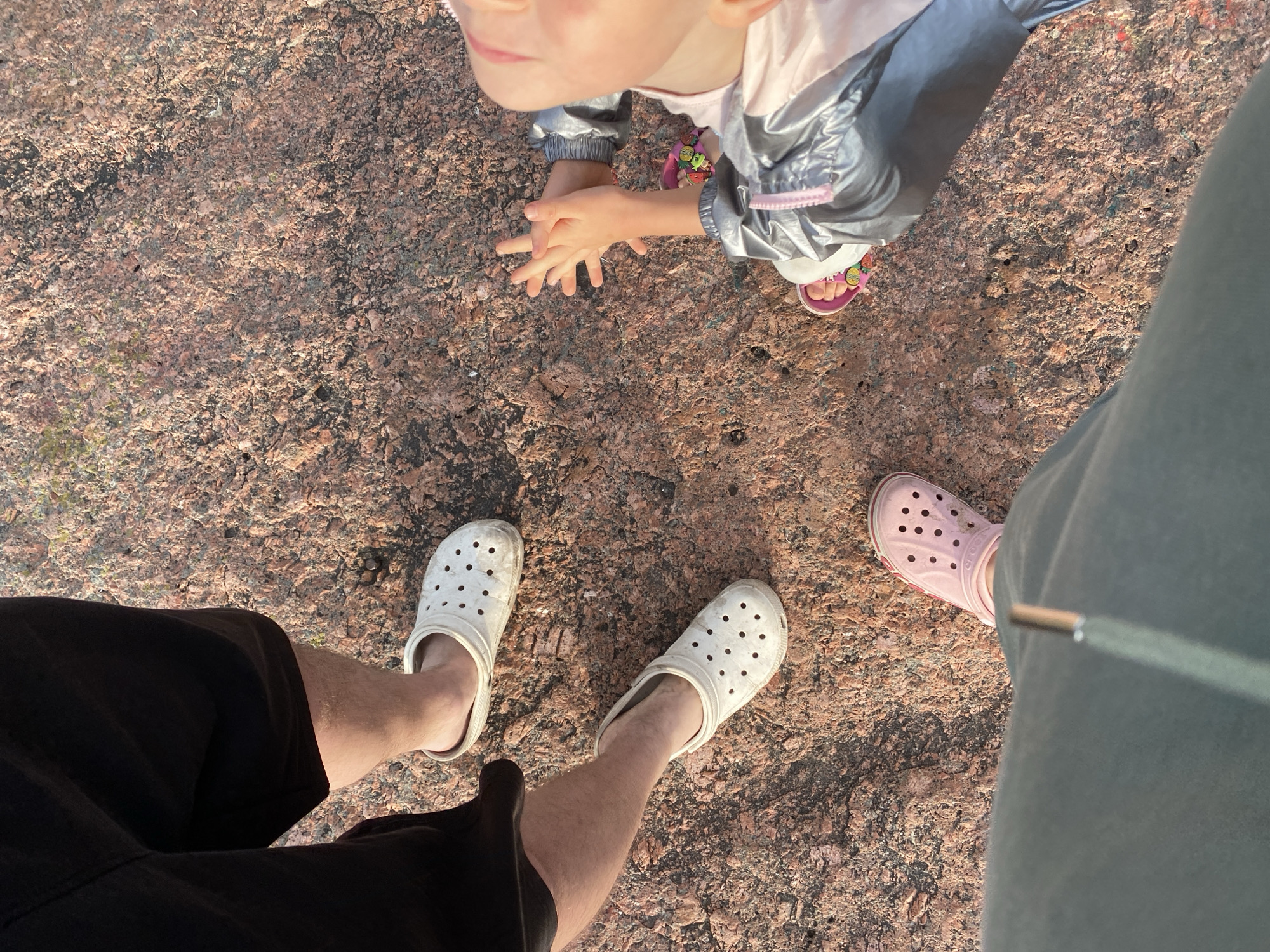 A group of people, including at least one adult and one child, stand on a large, reddish-brown rock surface. The adult is wearing black shorts and white Crocs. Another adult or older child is wearing dark pants and pink Crocs. The child is wearing a pink shirt and a silver jacket, and pink sandals with strawberry designs. The child's hands are clasped in front of them. The rock appears to be outdoors, with a textured, granular surface. The lighting suggests it is daytime, with sunlight illuminating the scene. The location is specified as Baltiiskoe, Russia.