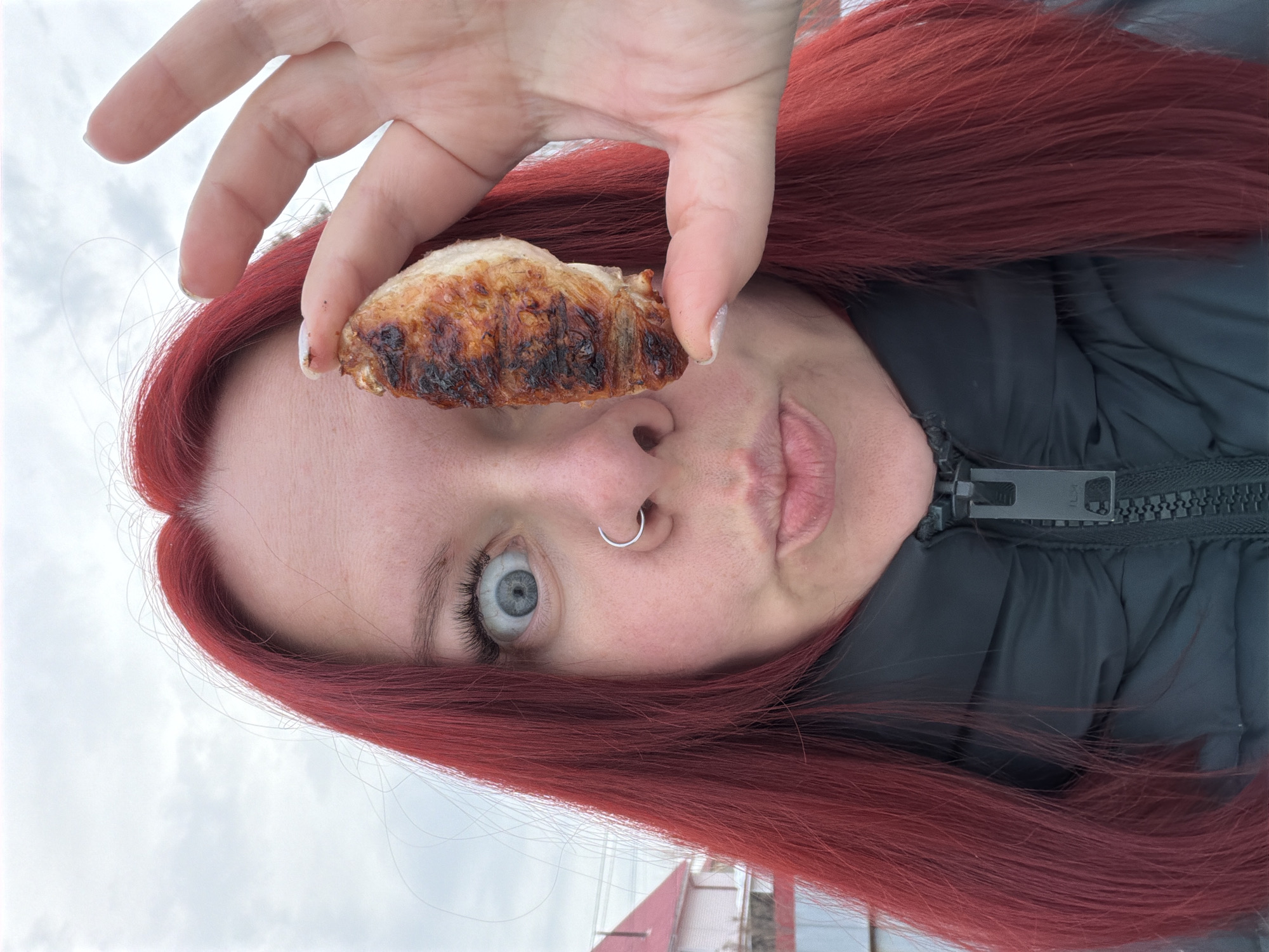 A frontal, close-up shot features an individual with vibrant red hair, fair skin, and a silver ring piercing the left nostril. The person wears a dark, possibly black, zipped puffer jacket. Their left eye is wide open with a visible grey-blue iris and pronounced eyelashes, while their lips are pursed. A piece of grilled meat, exhibiting browned and charred surfaces, is held vertically between the thumb and index finger of their right hand, deliberately obscuring their right eye. The background consists of an overcast, light-grey sky. Partially visible behind the individual's left side are a light-colored building with a red sloped roof and a dark fence or barrier. The scene is set outdoors in Chaikovsky, Russia.